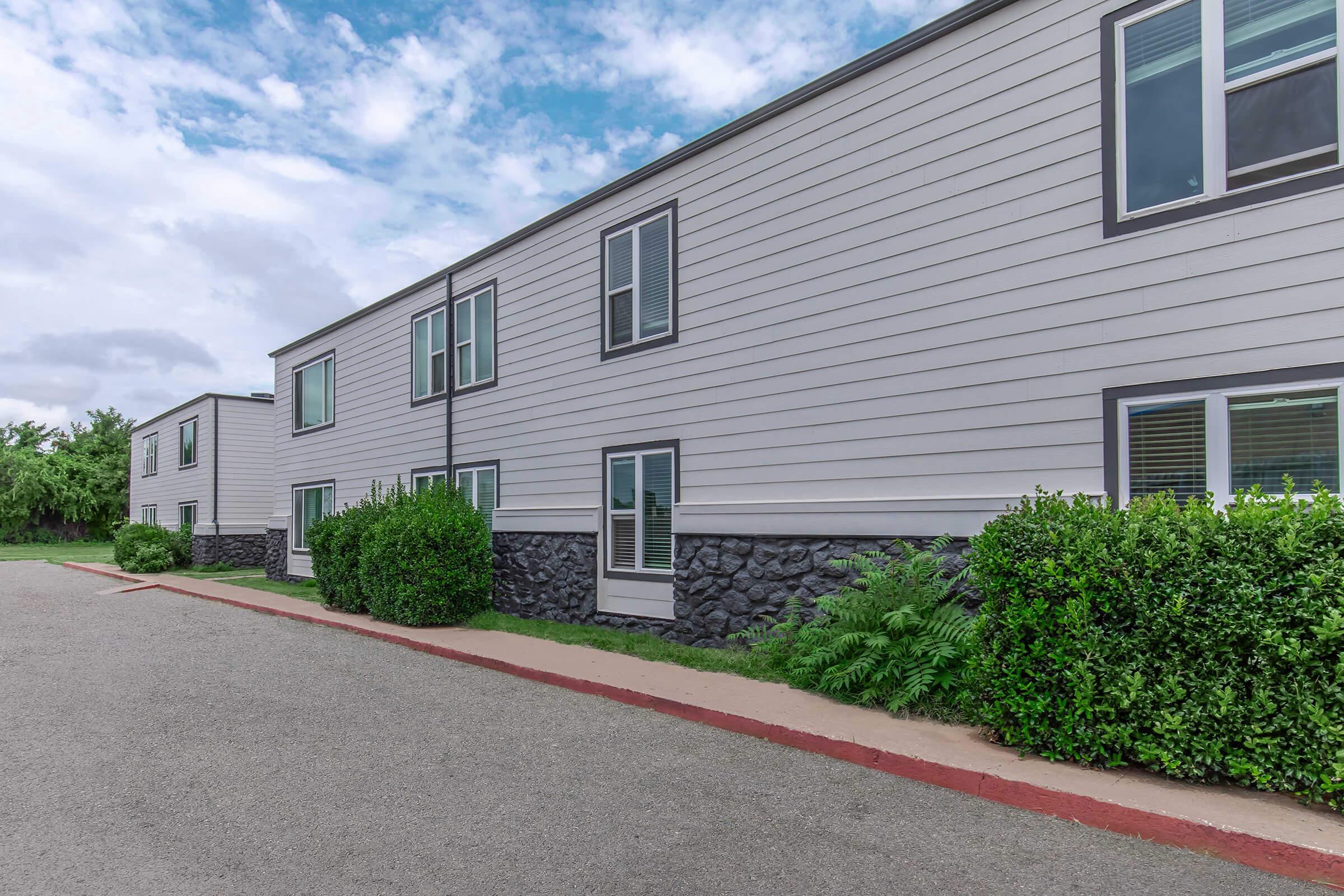 A side view of a modern building with light-colored siding and stone accents. The structure features multiple windows, surrounded by neatly trimmed green bushes. A paved path runs alongside the building, and the sky is partly cloudy.