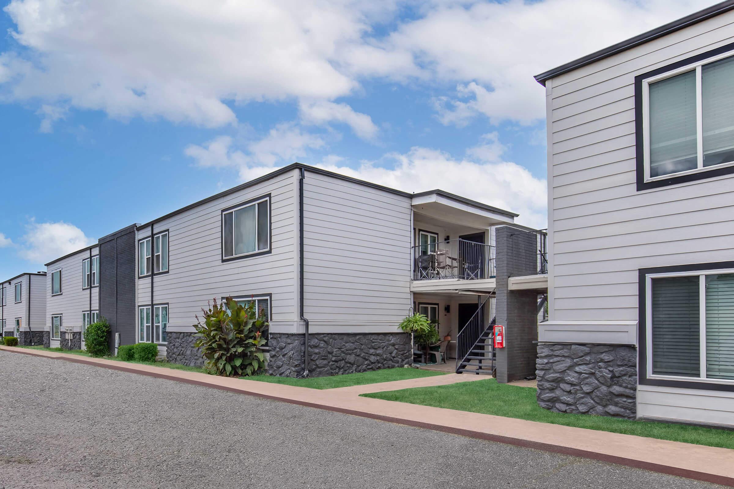 A row of modern, two-story residential buildings with a mixture of grey exterior and stone accents. There is a walkway leading to an entrance, with green plants and a light blue sky in the background. The scene conveys a clean and contemporary atmosphere in a community setting.