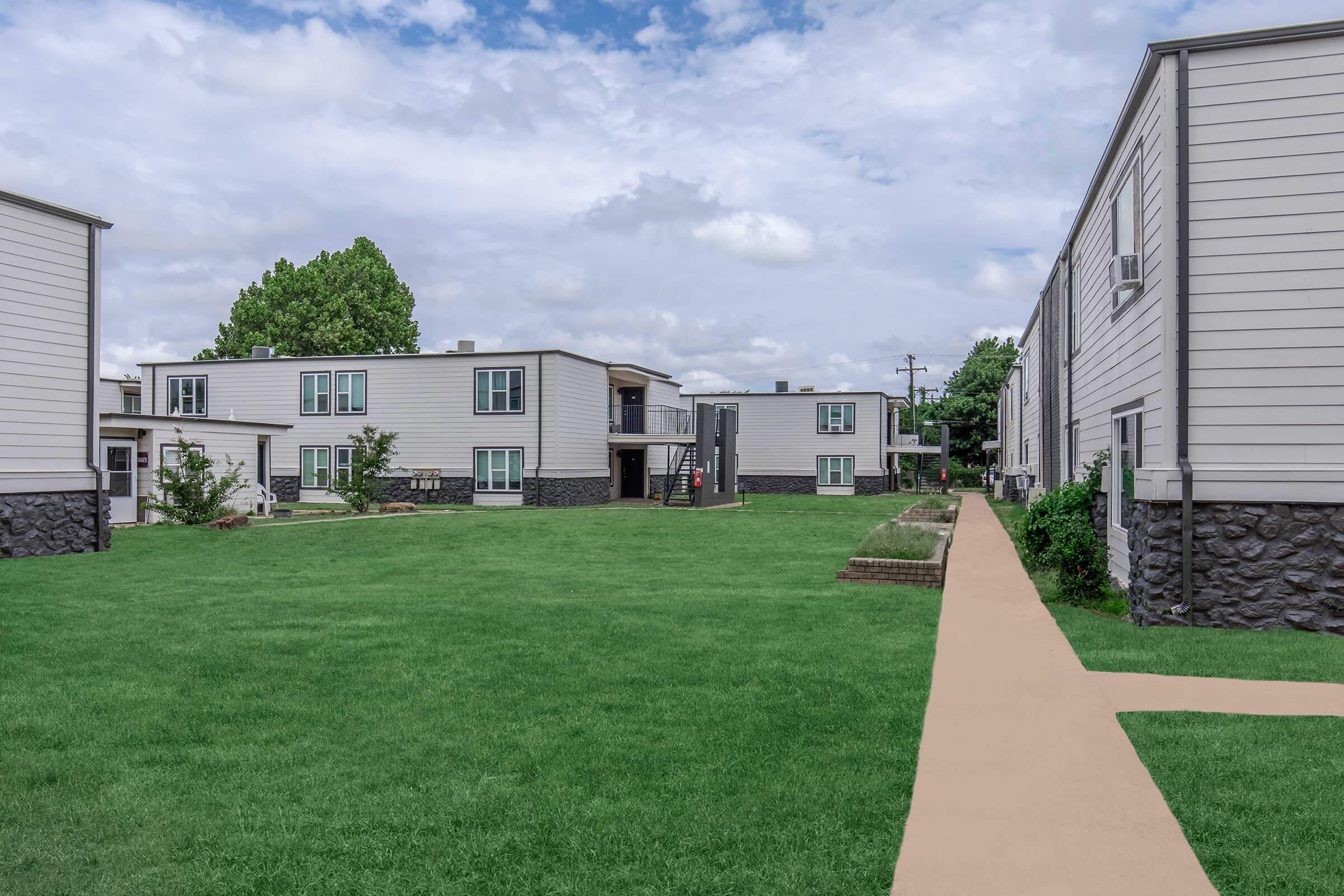 A view of a residential complex featuring multiple two-story buildings with light-colored siding and stone foundations. The foreground includes a well-manicured lawn, with a walkway leading through the grass. The sky is partly cloudy, creating a bright and inviting atmosphere.