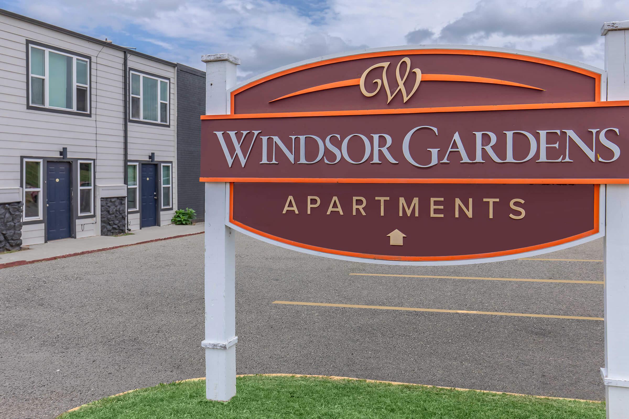 Sign for Windsor Gardens Apartments, featuring the name prominently with an arrow pointing upwards. In the background, there are modern apartment buildings with multiple doors and windows. The setting is outdoors with cloudy skies and a paved parking area.