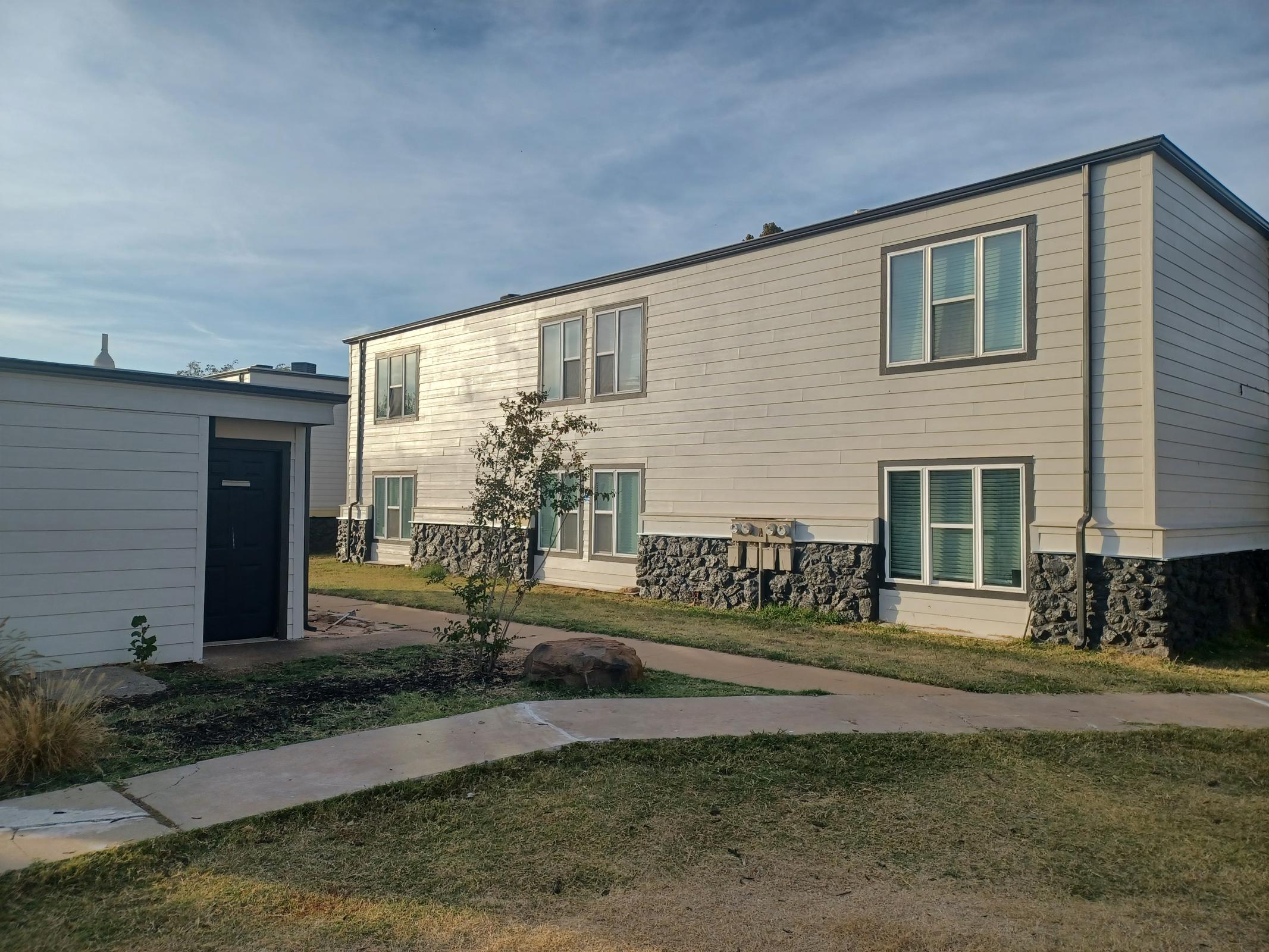 A residential building with two stories, featuring several windows and a stone foundation. There is a small grass area and a pathway leading to the entrance. A separate one-story structure is visible on the left, likely a mail area or storage. The sky is clear with a hint of clouds.