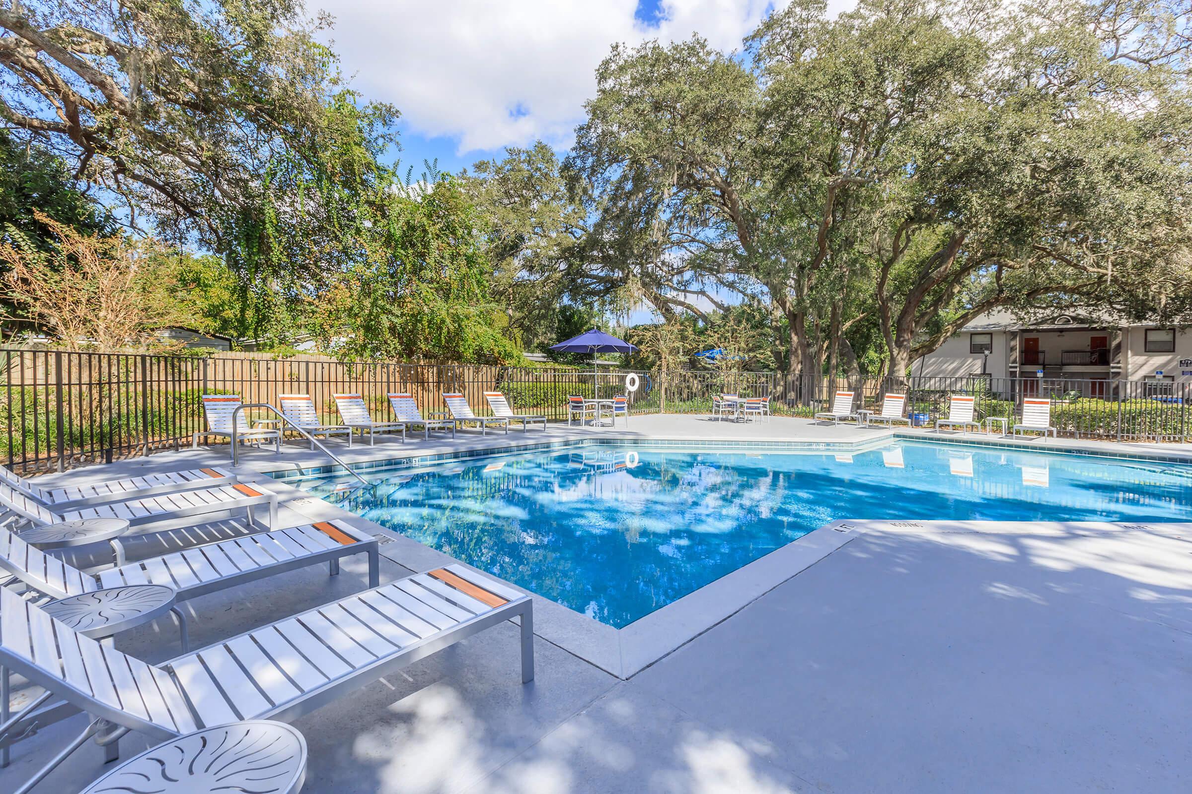A clean outdoor swimming pool surrounded by lounge chairs and shaded by trees. The pool area is enclosed by a fence, with umbrellas in the background. Bright blue sky with a few clouds enhances the inviting atmosphere of the space.