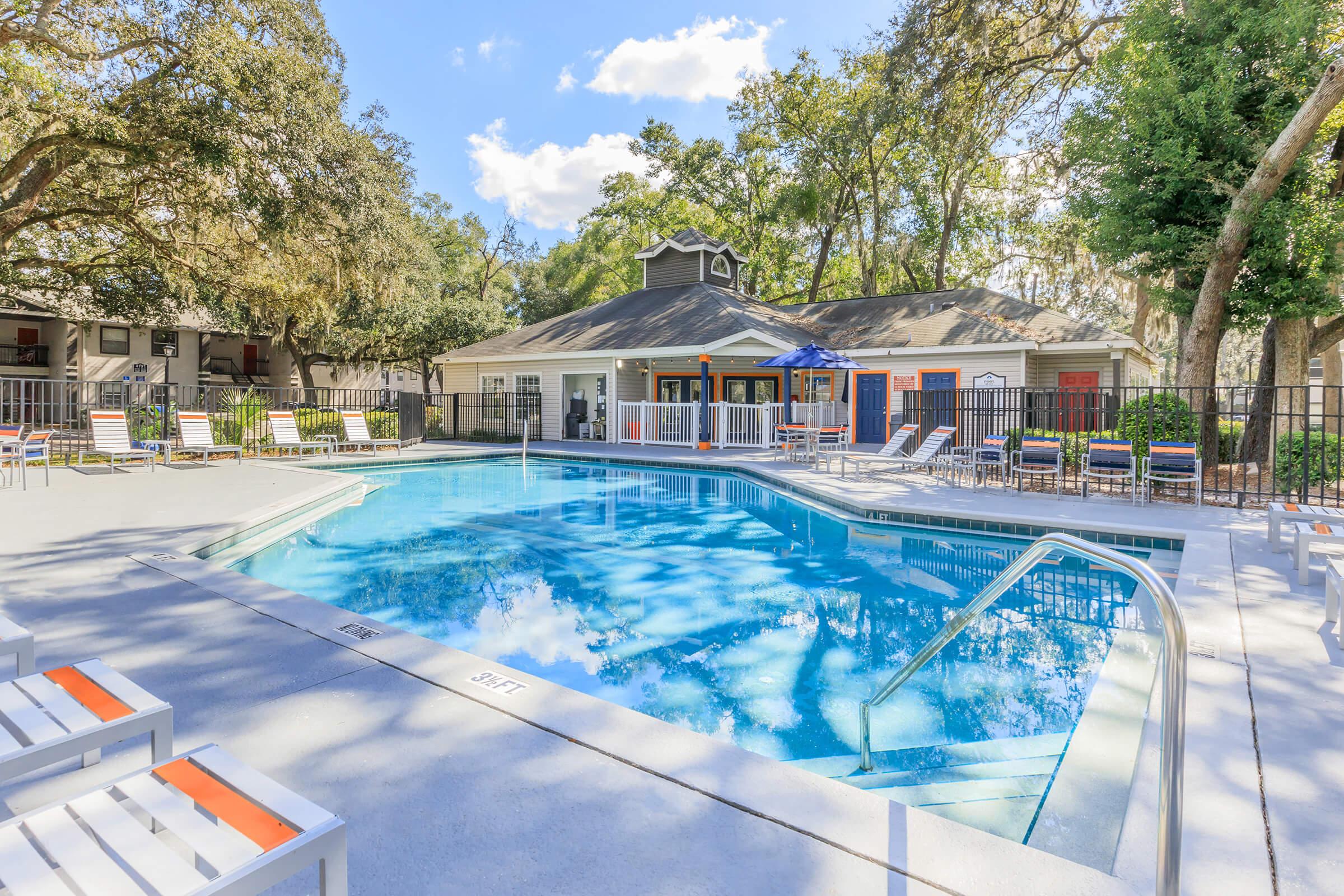 A serene outdoor pool area surrounded by lounge chairs and trees. The pool is clear and inviting, with a nearby clubhouse featuring large windows. The sky is partly cloudy, creating a peaceful atmosphere ideal for relaxation and leisure.