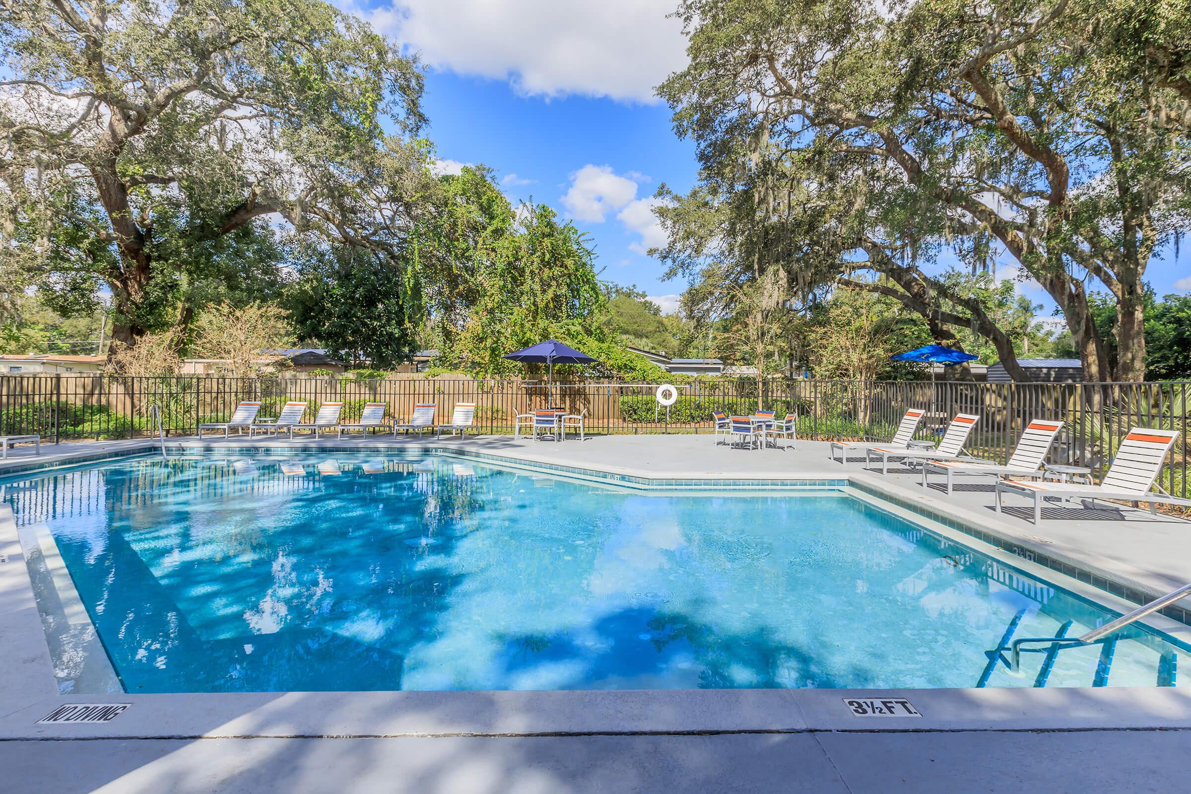 A bright outdoor swimming pool surrounded by lounge chairs and shaded by trees. The pool area features clear blue water and a sunny sky with fluffy clouds, creating a relaxing atmosphere. Umbrellas are set up for shade, making it an inviting space for leisure and relaxation.
