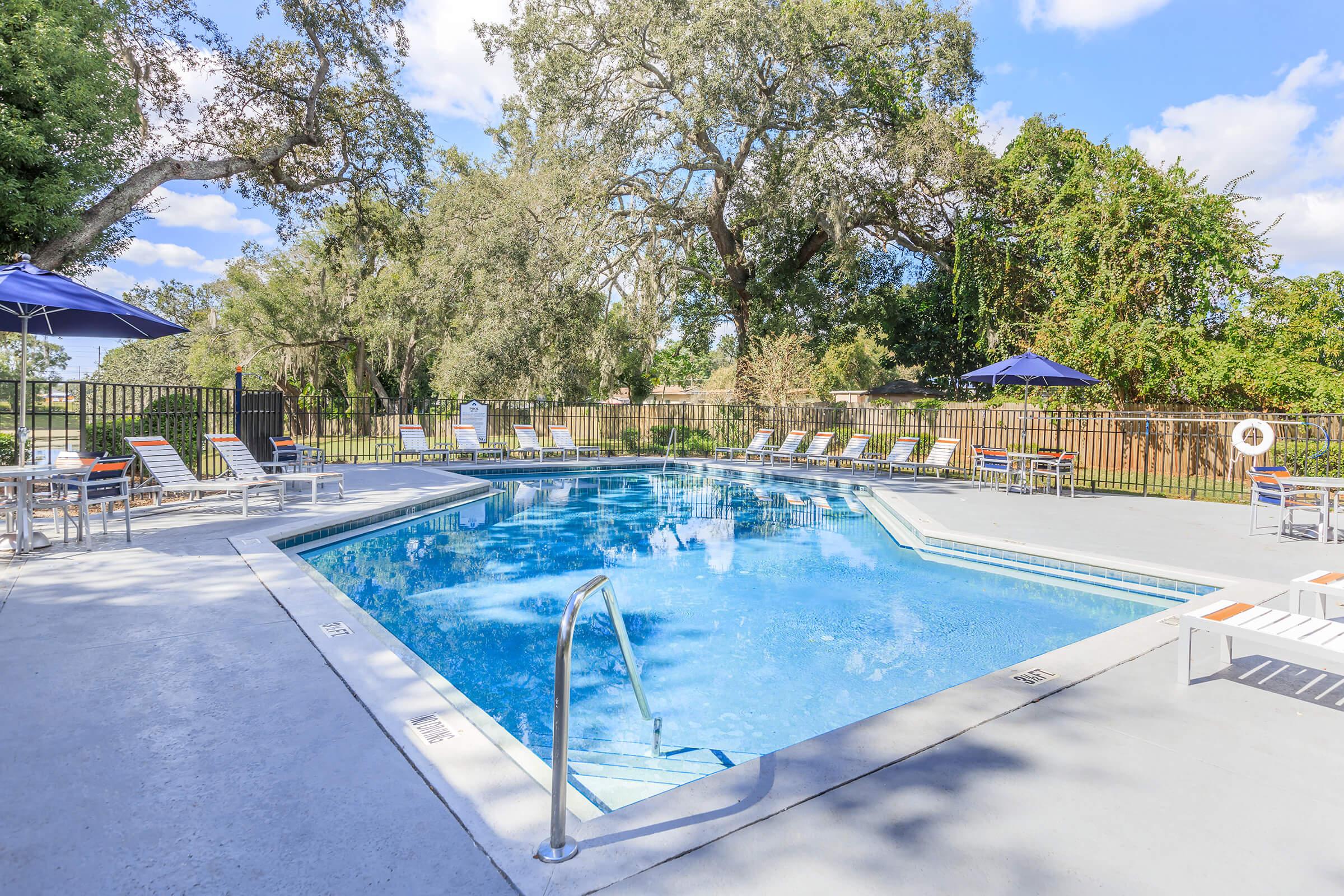 A clear blue swimming pool surrounded by a landscaped area with trees and a fence. There are lounge chairs and umbrellas positioned around the pool, providing a relaxing outdoor atmosphere. The sky is partly cloudy, adding to the serene setting.