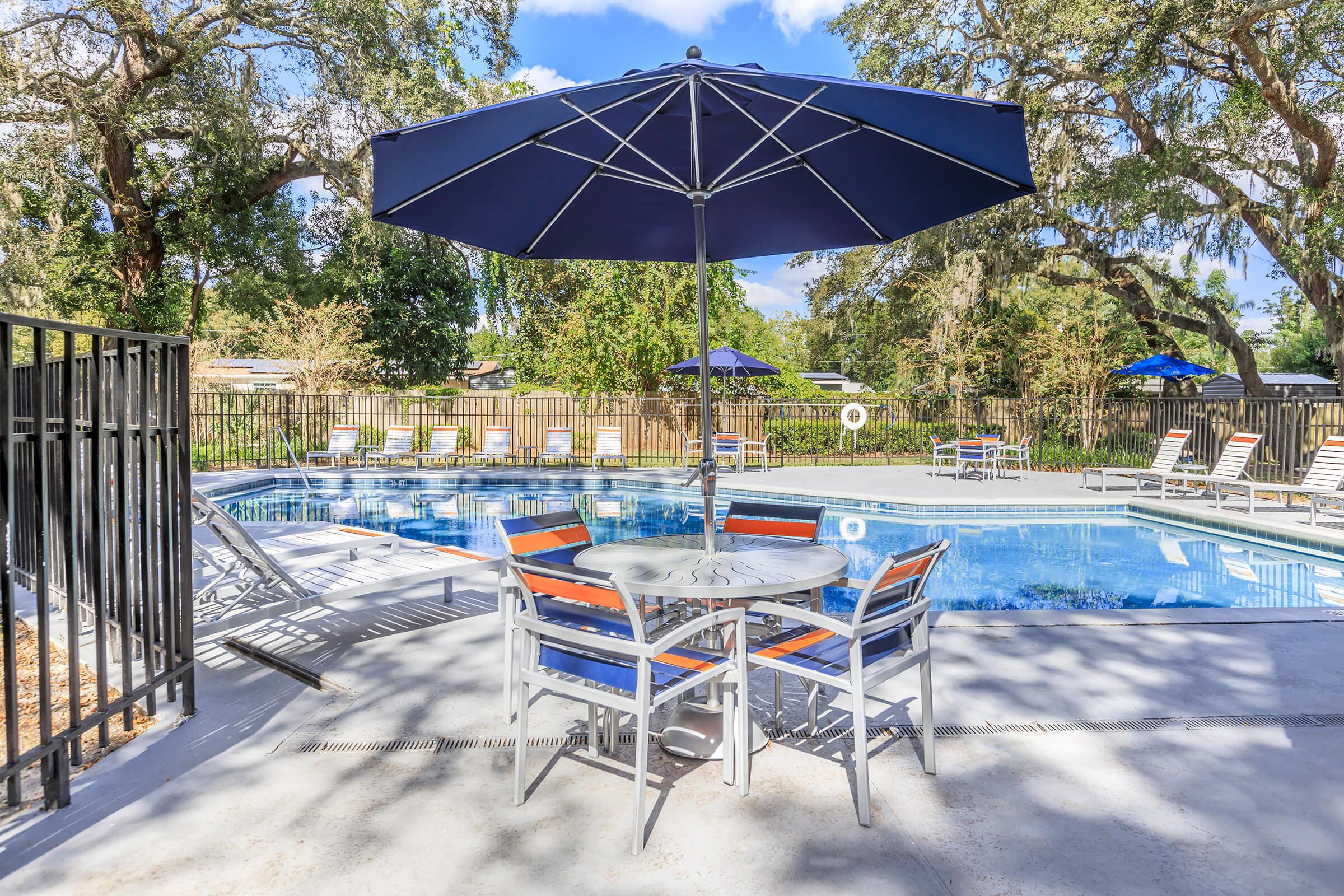 A relaxing outdoor pool area featuring a blue umbrella shaded table and chairs, surrounded by lounge chairs. The pool is clear and inviting, with green trees in the background and a fenced perimeter, creating a tranquil atmosphere for leisure and sunbathing.