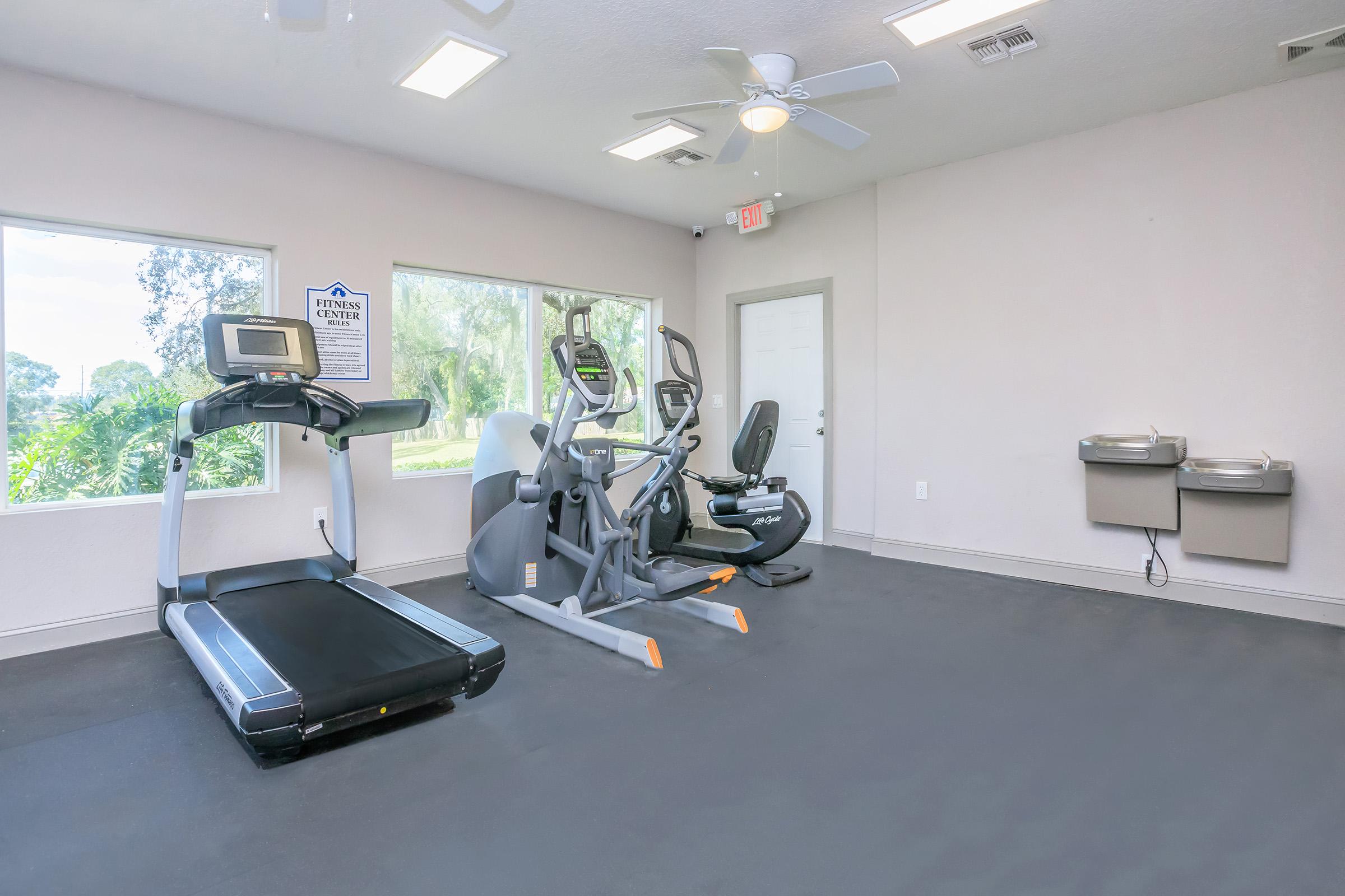 A bright, modern fitness room featuring a treadmill and an elliptical machine, with large windows allowing natural light. There are two water fountains on the wall, and the space has a clean, minimalist design with light-colored walls and a dark floor.