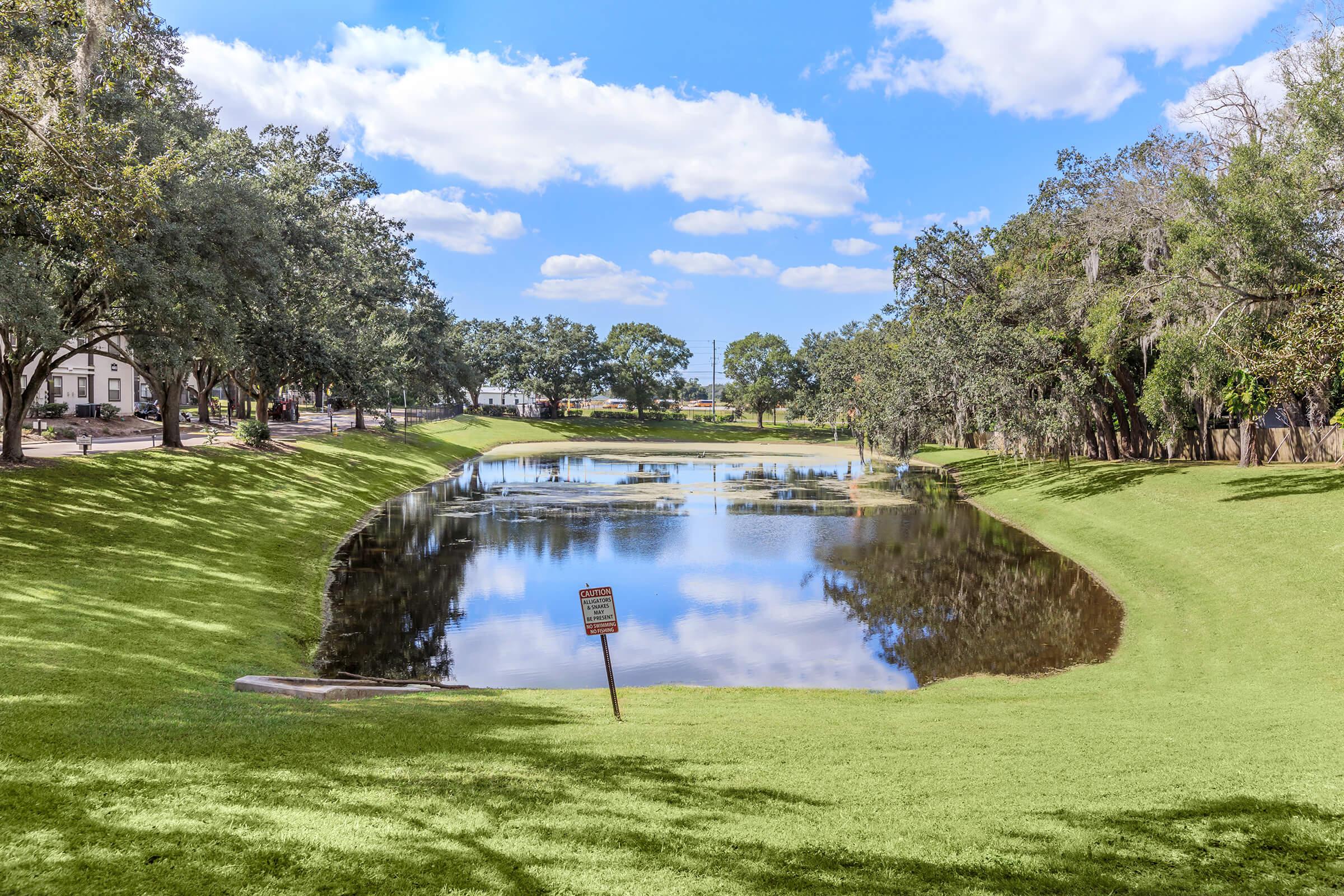 A peaceful park scene featuring a calm pond surrounded by lush green grass and trees. A sign is visible near the water's edge, and the sky is partly cloudy. The reflection of the trees and clouds can be seen in the water, creating a serene atmosphere.