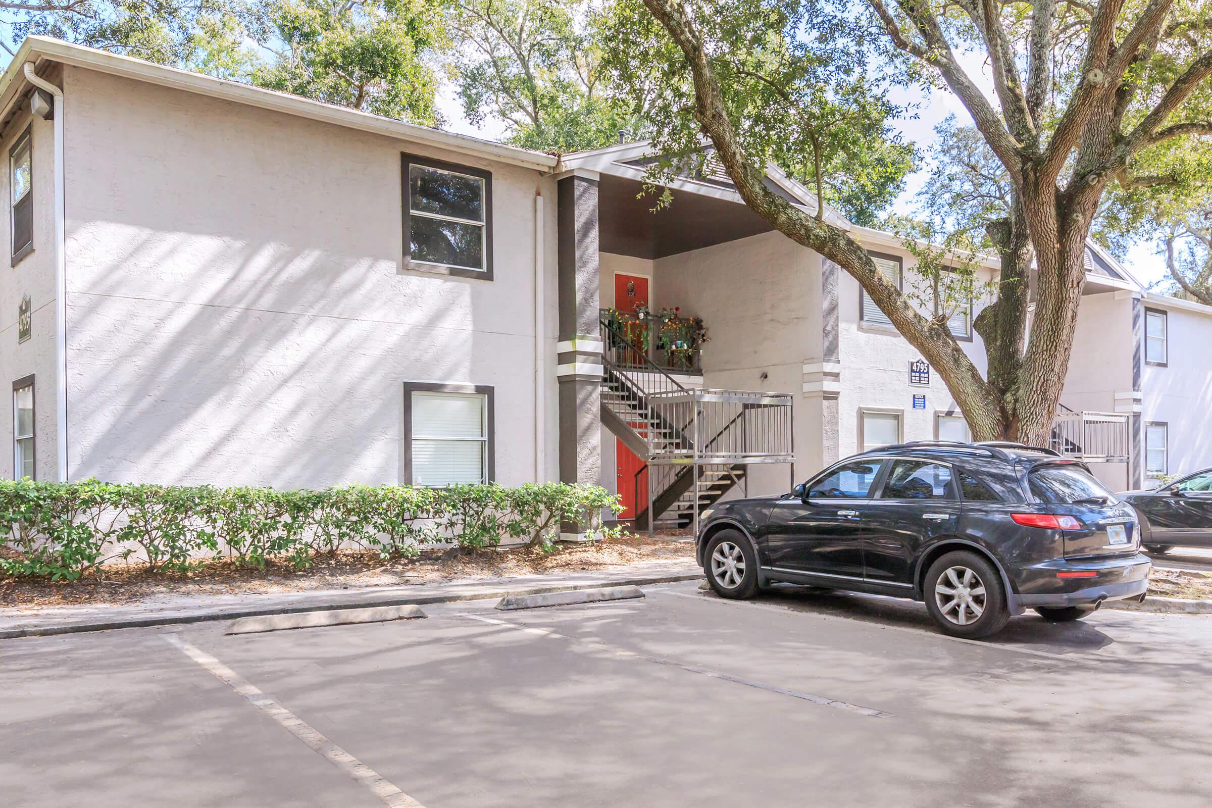Two-story apartment building with a gray exterior and orange accent door. A staircase leads to the second floor, decorated with plants on the landing. A black car is parked in the foreground, with additional parking visible. Lush greenery surrounds the building, providing a natural setting.