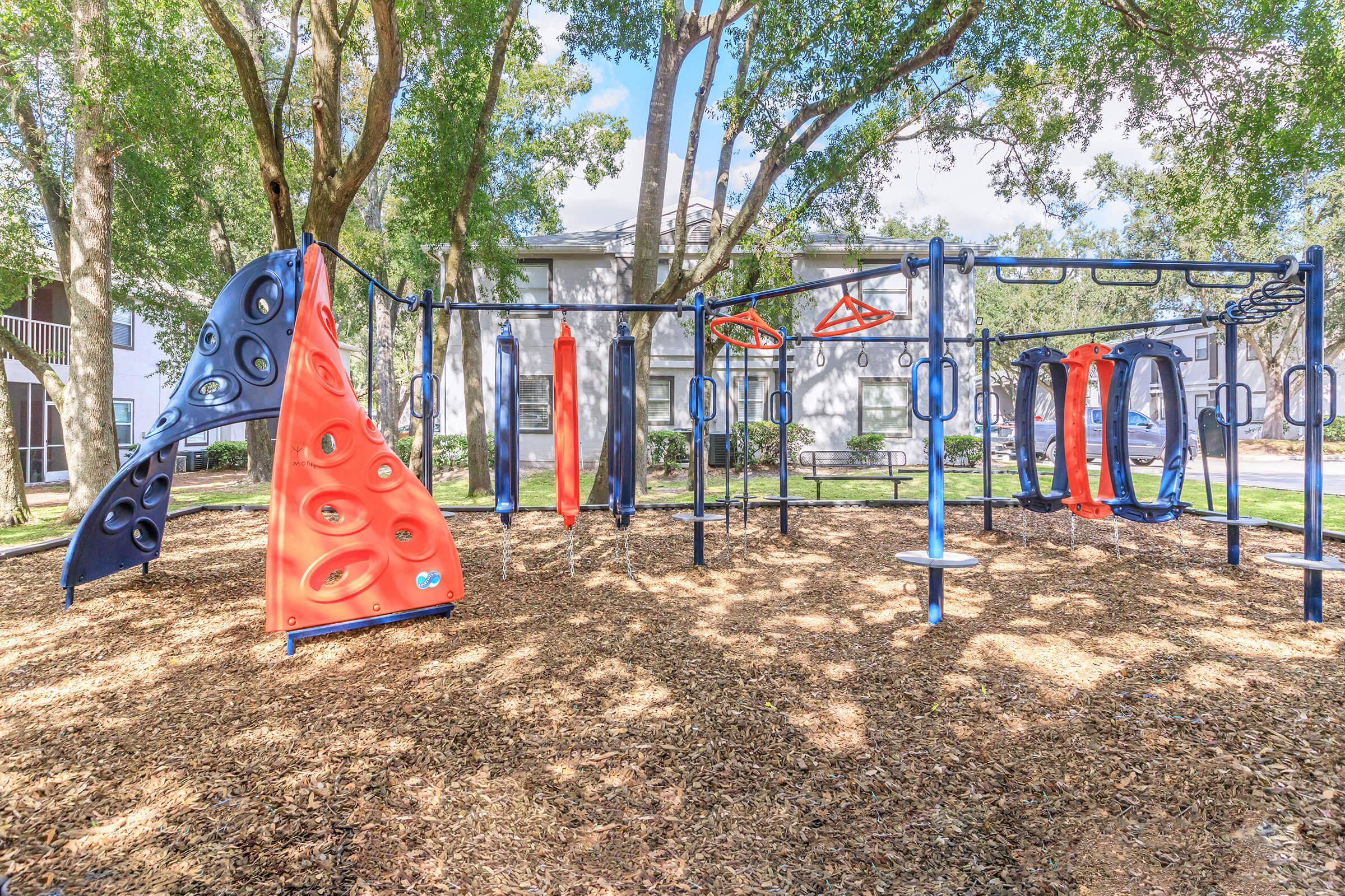 Colorful playground equipment in a park setting, featuring climbing structures, swings, and slides. Surrounded by trees and a residential building in the background. Soft ground covered with wood chips for safety. Bright, sunny day with scattered clouds.