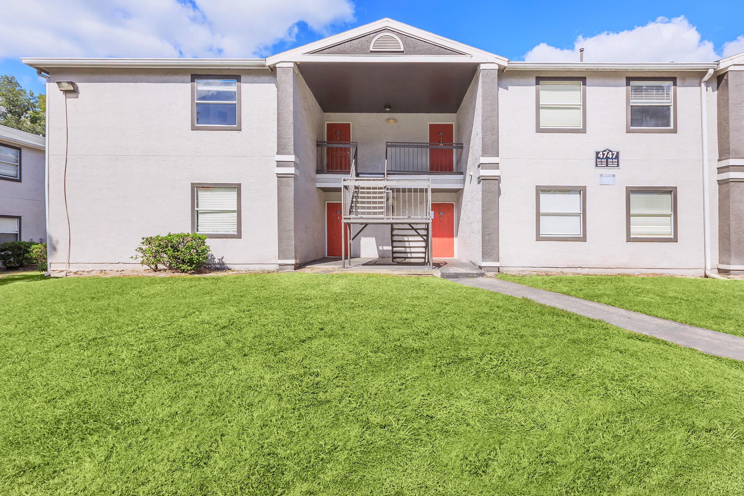 Exterior view of a two-story apartment building with gray walls and red doors. A staircase leads to the second floor in the middle, surrounded by well-maintained green grass. The sky is partly cloudy, adding a bright atmosphere to the scene.
