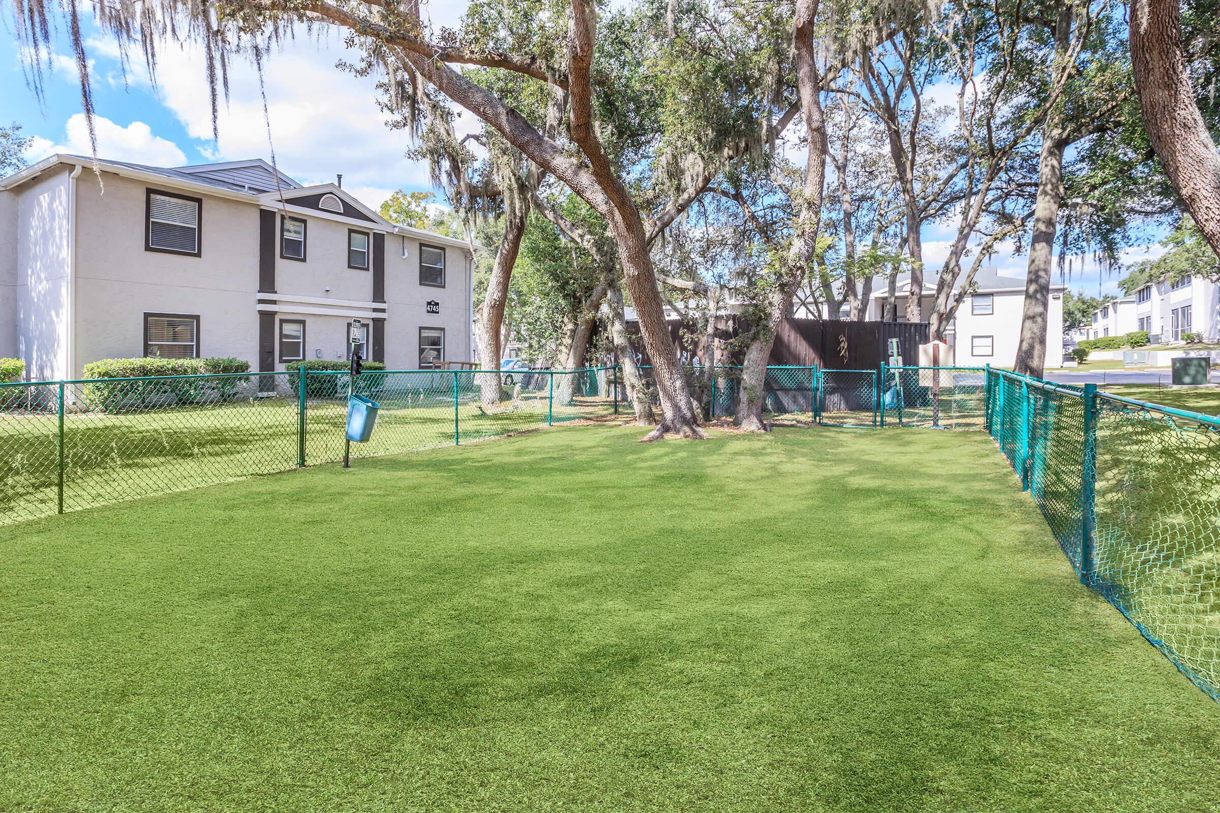 A well-maintained grassy area enclosed by a blue fence, with a couple of trees providing shade. In the background, a two-story building and another structure are visible under a bright blue sky with scattered clouds. The scene is tranquil and inviting for outdoor activities.