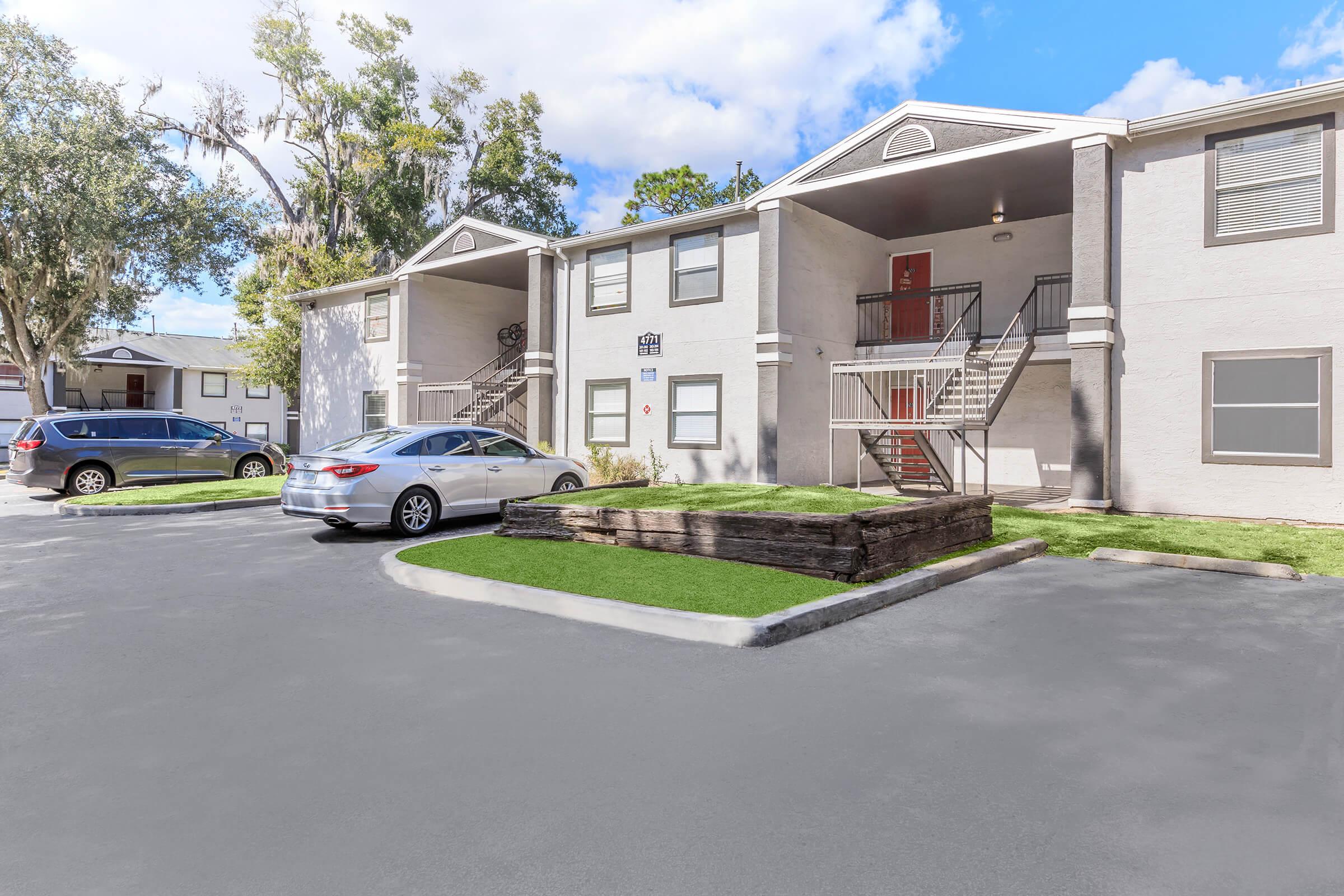 Exterior view of a two-story apartment building with a light-colored facade. The entrance has stairs leading to the upper floor. In the foreground, there are two parked cars, and a small patch of grass with wooden landscaping. The sky is partly cloudy, contributing to a bright atmosphere.