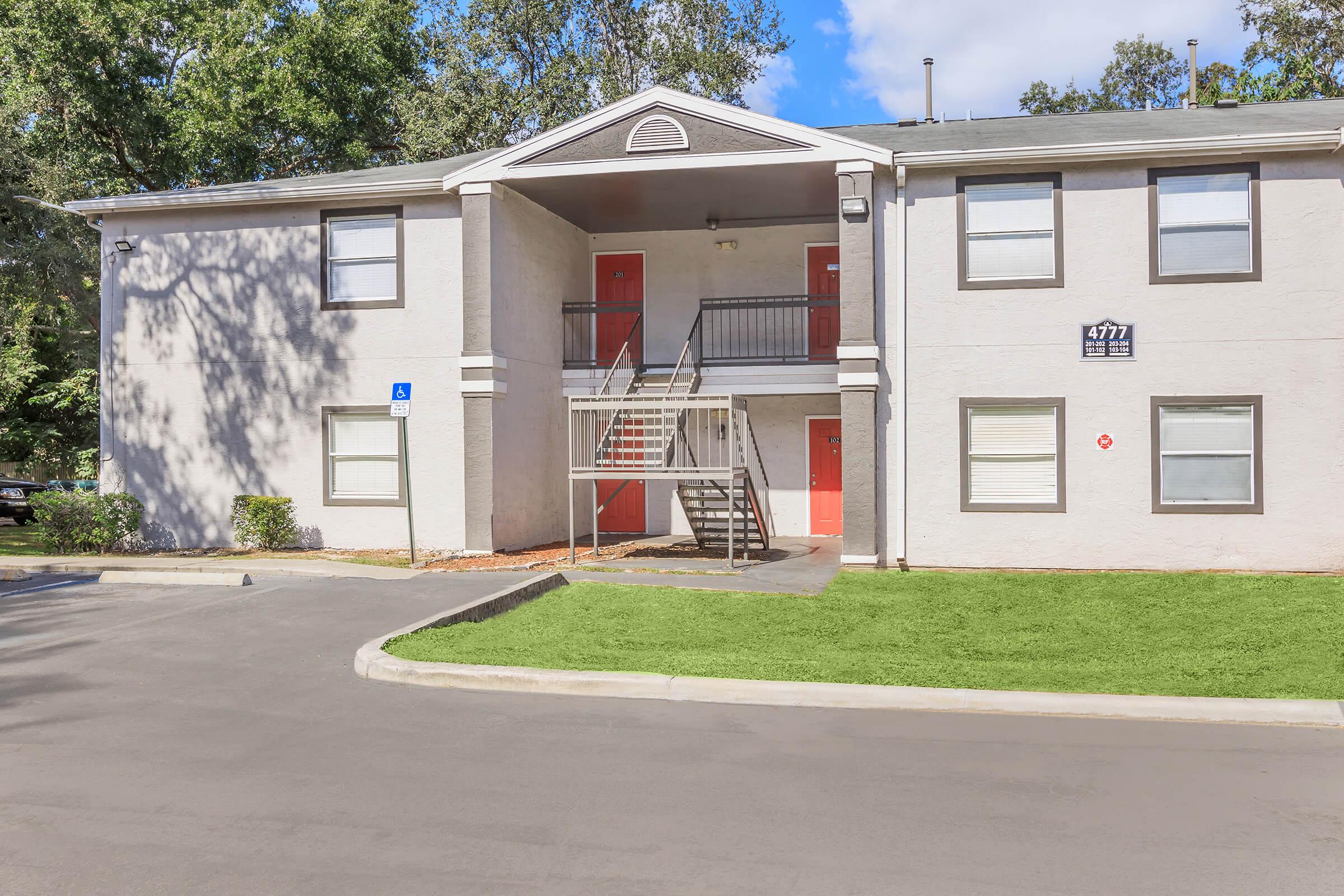 A two-story apartment building with light-colored exterior walls. The entrance has a set of stairs leading to a second-floor porch, flanked by two red doors. There is a well-maintained green lawn in front of the building, with a paved parking area nearby. Trees provide some shade around the property.