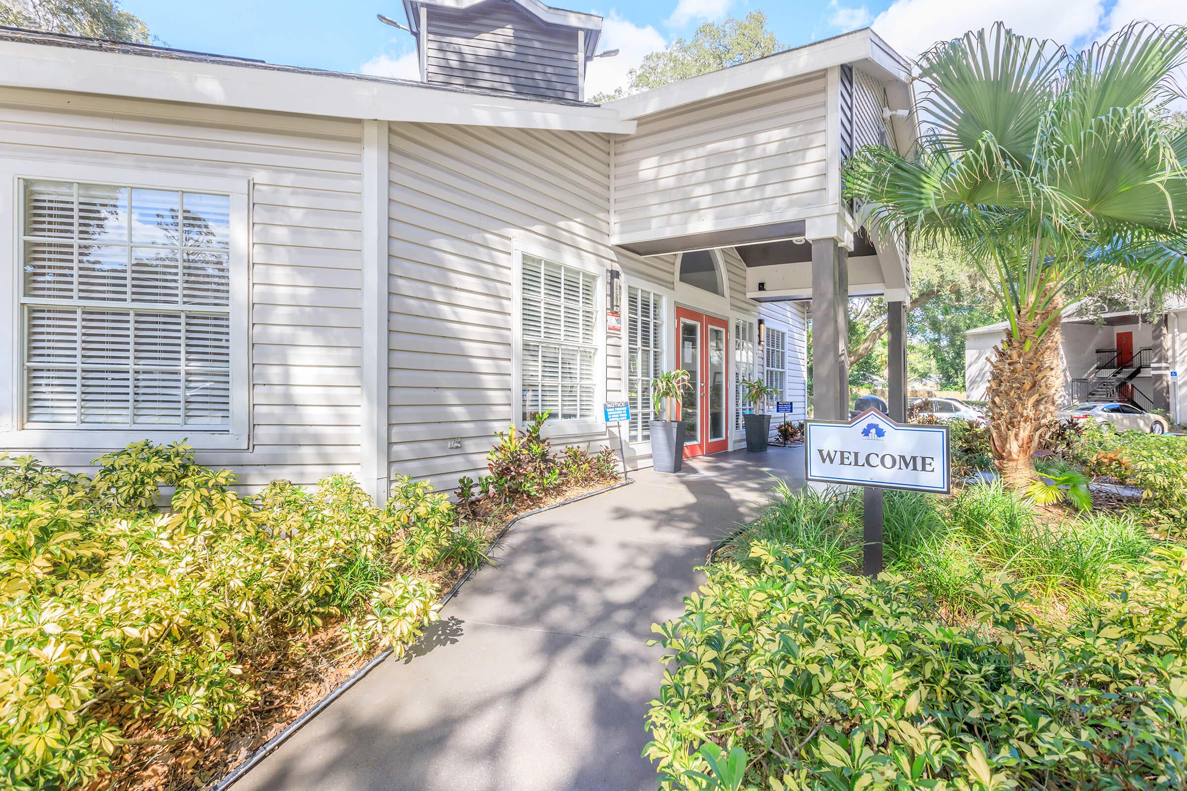 A welcoming entrance to a building with a sign that reads "WELCOME." The path is lined with green plants and shrubs, and there are large windows on the facade. The architecture features light-colored siding and a gabled roof, providing a bright and inviting atmosphere.