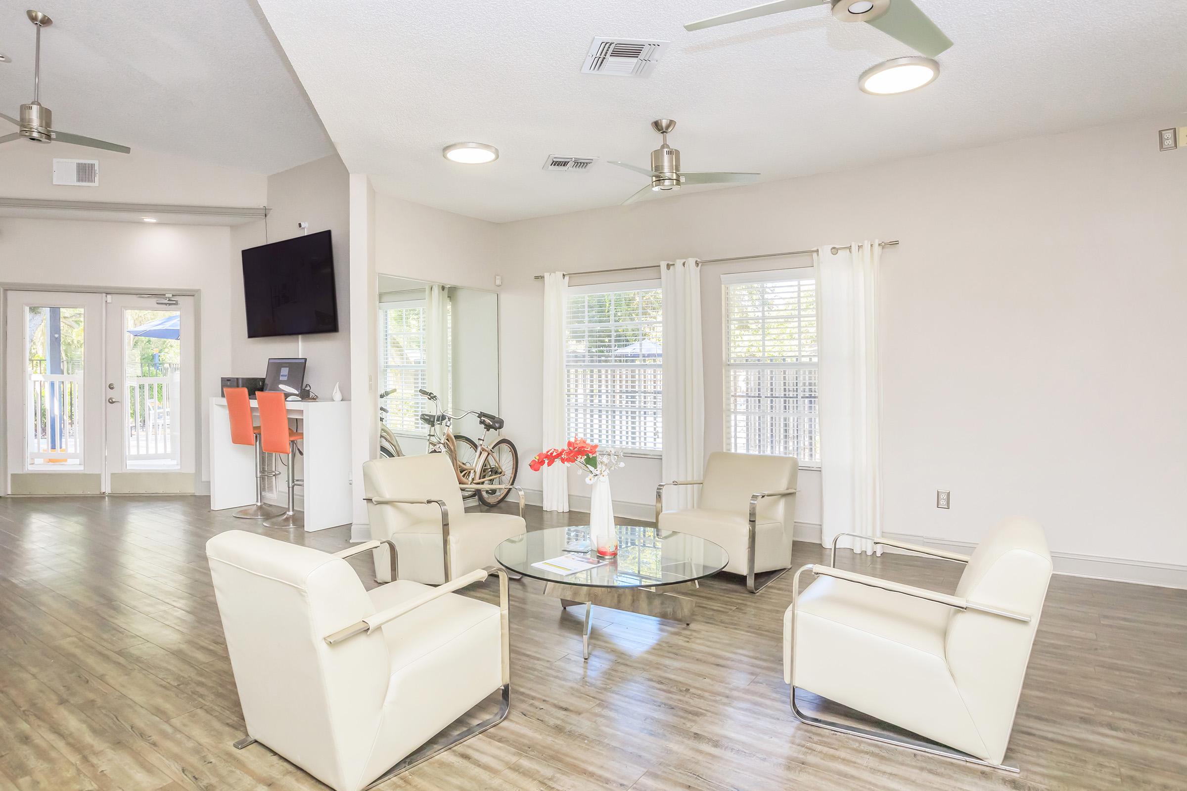 A modern lounge area featuring white armchairs arranged around a glass coffee table. There are large windows with white curtains allowing natural light, a television on the wall, and a bike visible in the background. The floor is a light wood finish, creating a bright and inviting atmosphere.