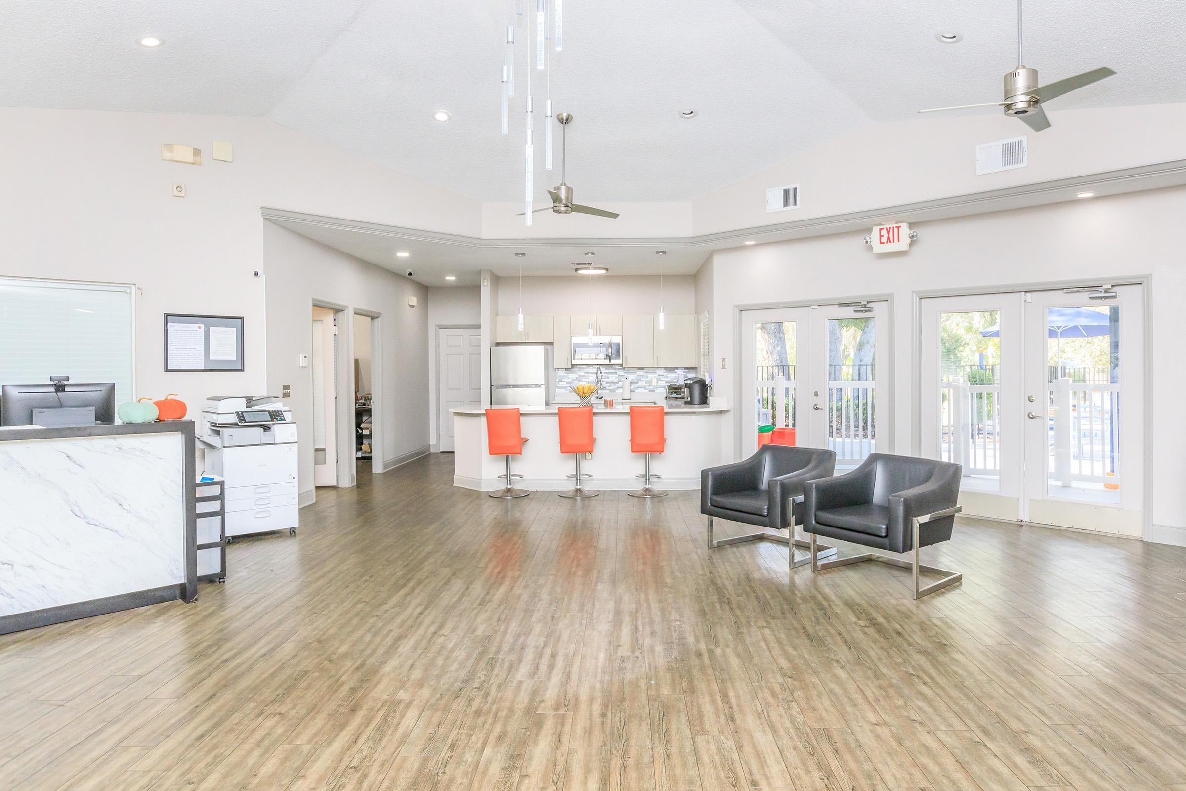 Modern lobby area with a reception desk, seating for visitors, and a kitchenette. The space features contemporary decor, including orange bar stools and sleek black chairs, with large windows allowing natural light to fill the room. The floor has a wood-like finish, contributing to a warm atmosphere.