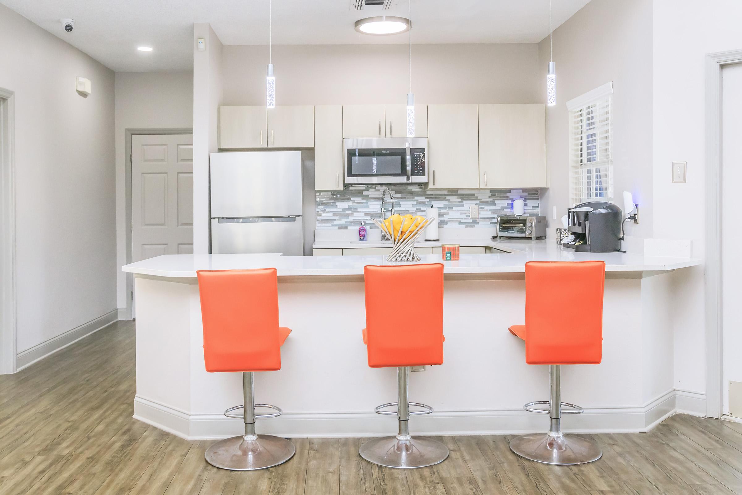 Modern kitchen featuring a white countertop with three orange bar stools, stainless steel refrigerator, microwave, and a sleek backsplash. Natural light illuminates the space, highlighting contemporary fixtures and decor, creating a bright and inviting atmosphere.