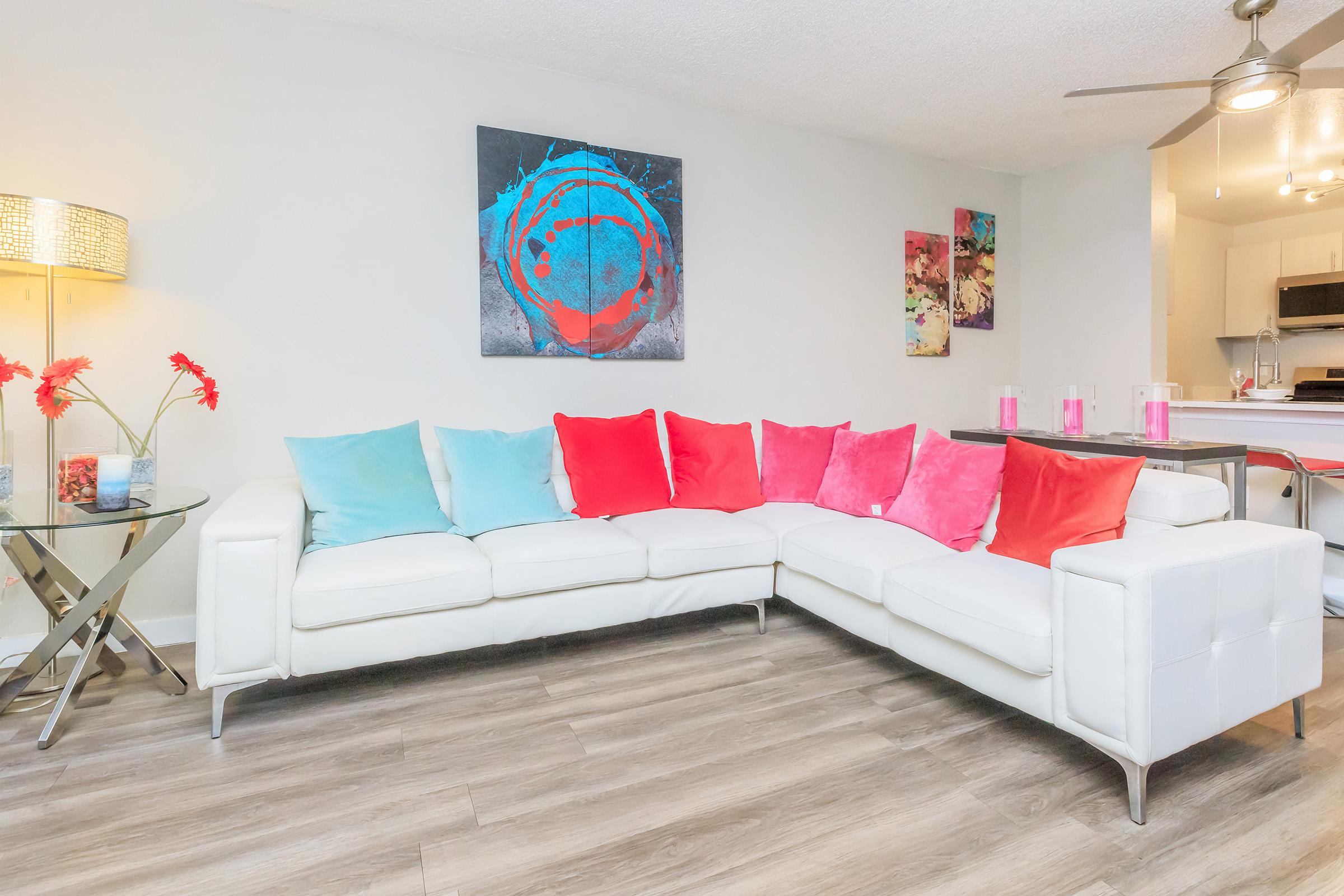 A modern living room featuring a white sectional sofa adorned with colorful cushions in shades of turquoise and pink. A stylish lamp with a decorative shade and a round table with a vase of red flowers are positioned nearby. A vibrant abstract painting hangs on the wall, adding a pop of color to the space.