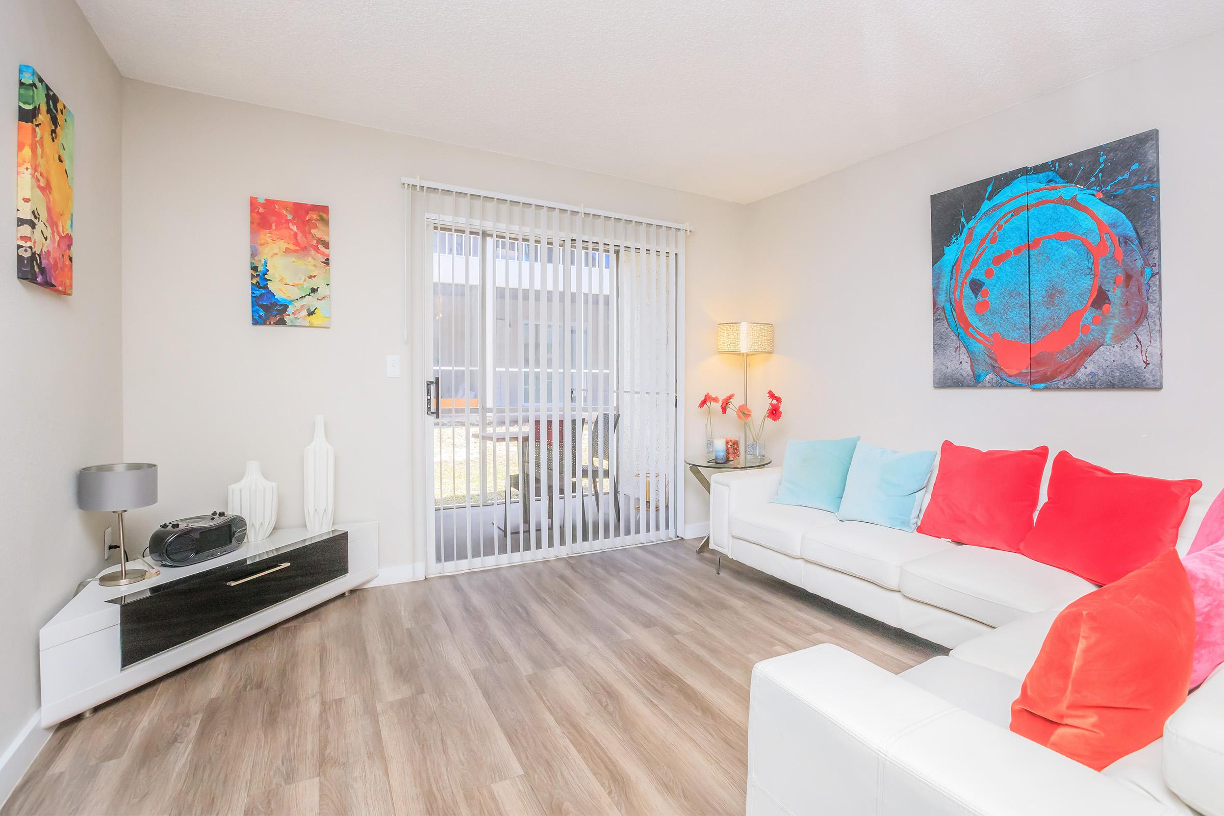 A modern living room featuring a white sofa with colorful cushions in shades of blue and orange. There are abstract paintings on the walls, a stylish lamp, and a decorative console. Large sliding doors lead to a balcony, and the floor has a warm wooden finish.