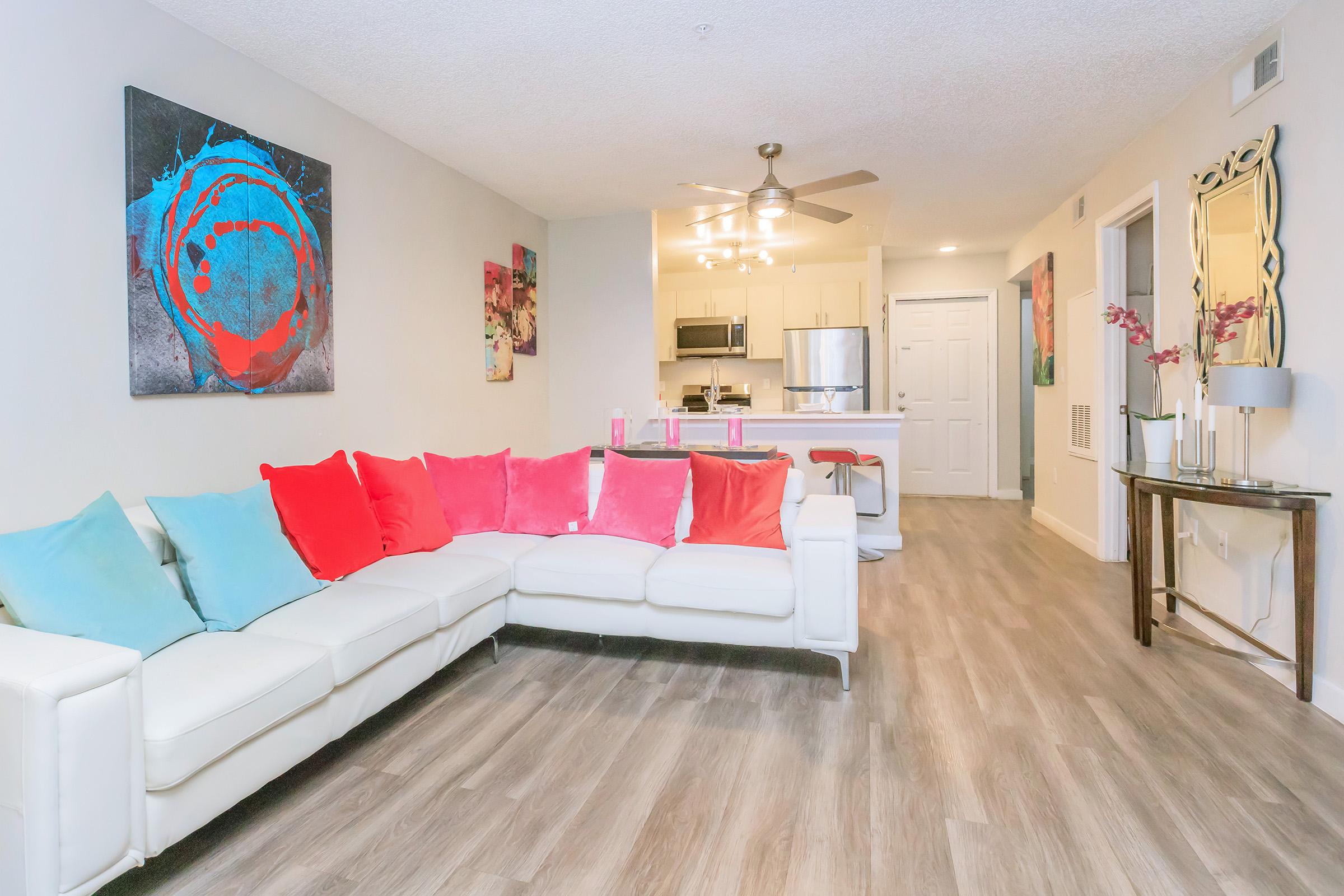 A modern, bright living room featuring a white sectional sofa with colorful pink and blue pillows. There are abstract paintings on the walls, a ceiling fan, and hardwood flooring. A dining area and kitchen are visible in the background, with a stylish mirror and decor on a side table.