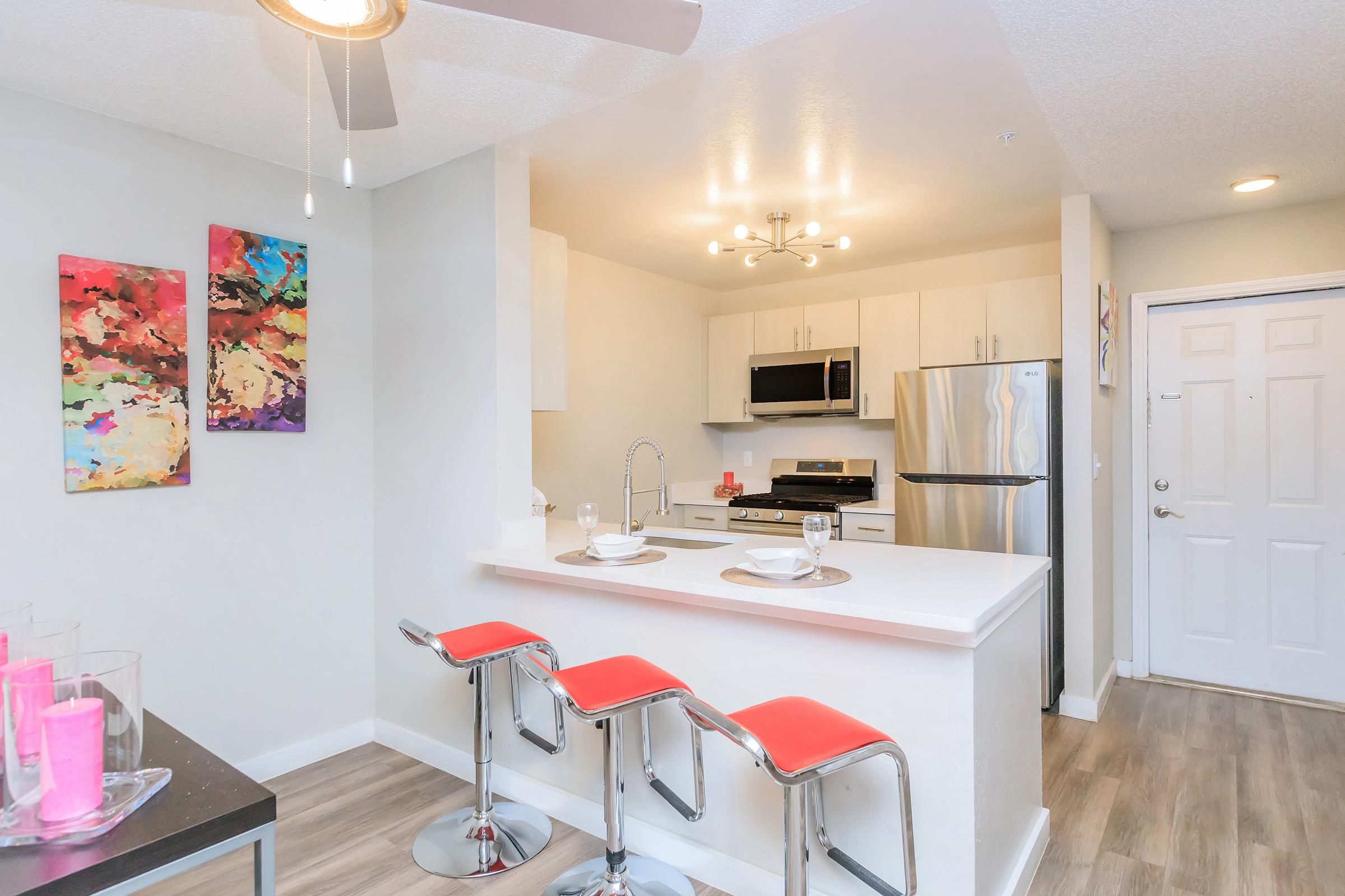 Modern kitchen with a white countertop and stainless steel appliances, featuring a dining bar with three red stools. The walls are painted in light colors, adorned with colorful artwork. A door leads outside, and the space is illuminated by a ceiling fan and natural light.