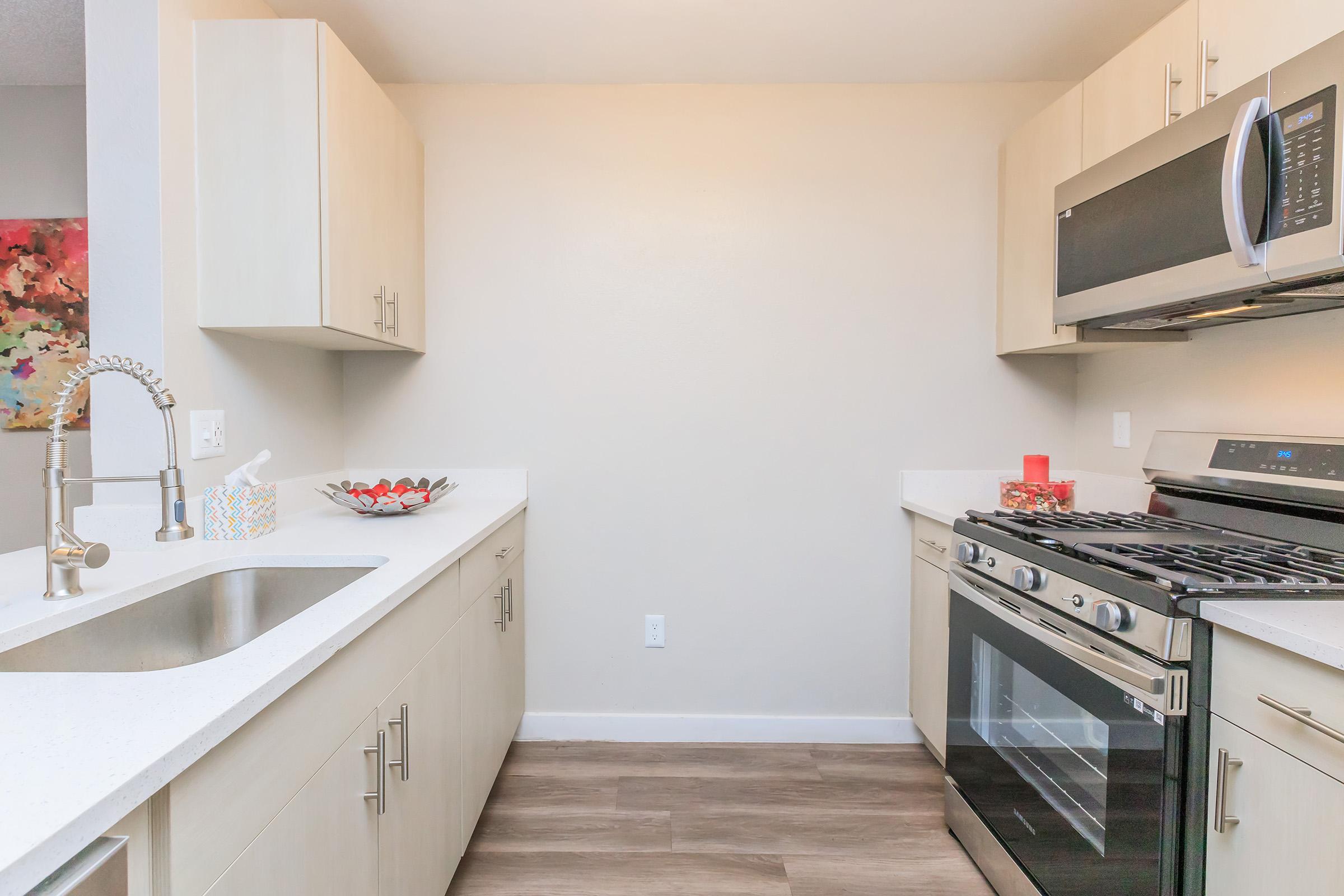 Modern kitchen with light-colored cabinets and a clean design. Features a stainless steel refrigerator, oven, and microwave. A white countertop with a sink and a bowl of fruit adds a pop of color. The walls are a neutral tone, creating a bright and spacious atmosphere. Wooden flooring completes the contemporary look.