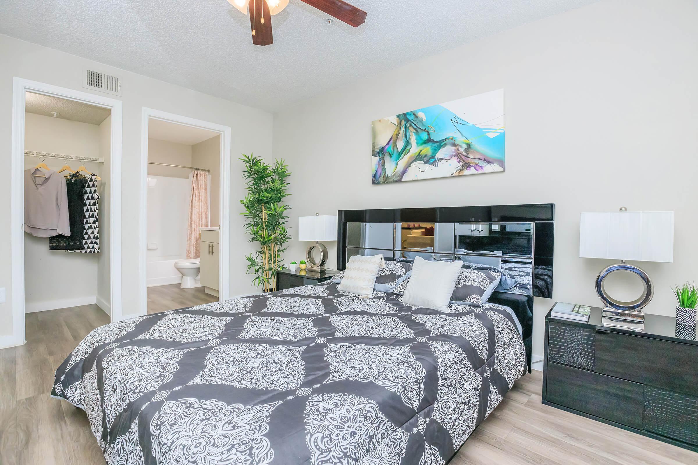 A modern bedroom featuring a large bed with a decorative black and white bedspread. There's a stylish headboard, nightstands with lamps on either side, and a piece of abstract art above the bed. A small plant and decorative items are on the nightstands. A closet and bathroom are visible in the background.