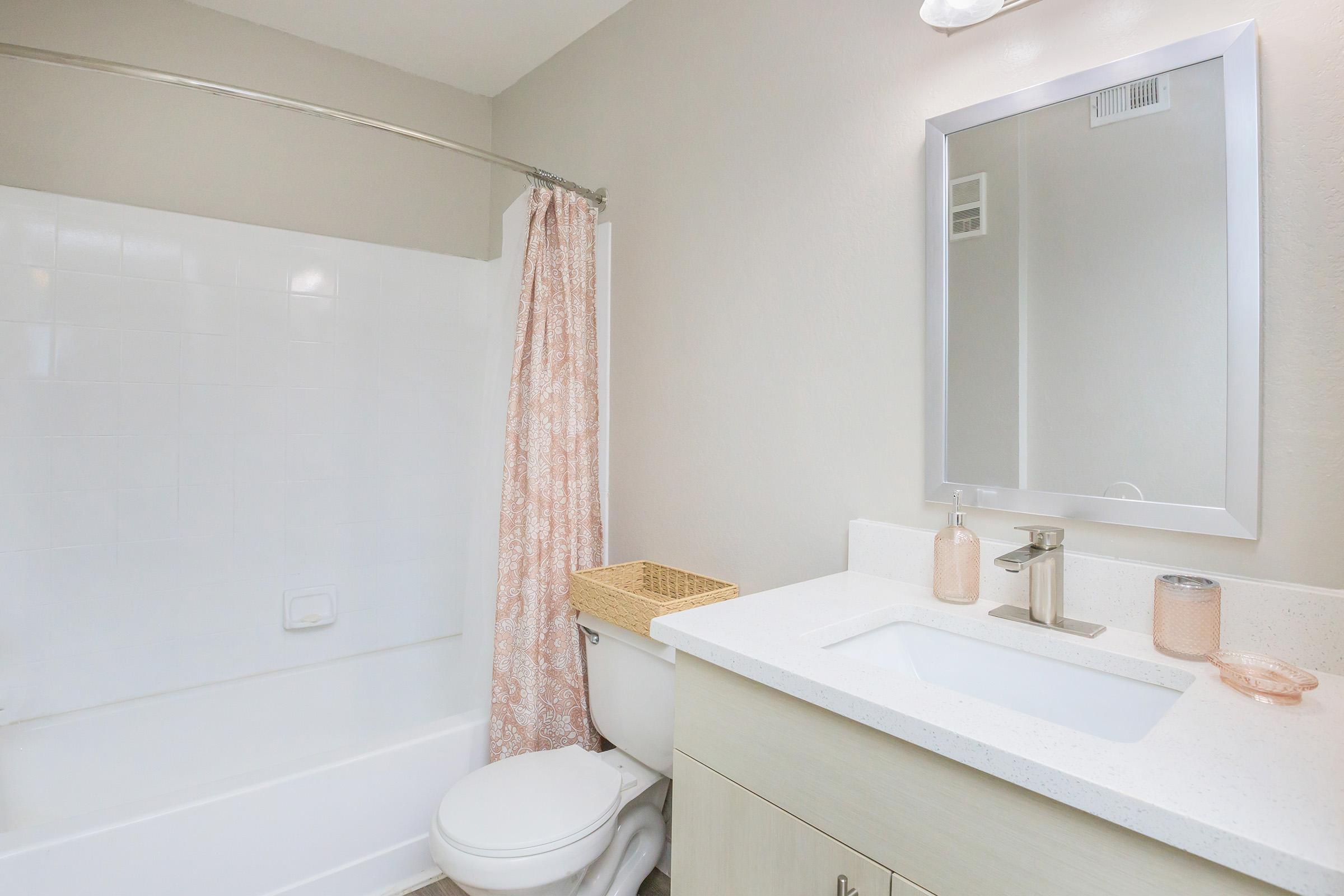 A modern bathroom featuring a white bathtub with a shower curtain, a wall-mounted mirror above a sink with a light-colored countertop, a beige cabinet, and decorative items in soft colors. The walls are painted in a light shade, creating a clean and contemporary look.
