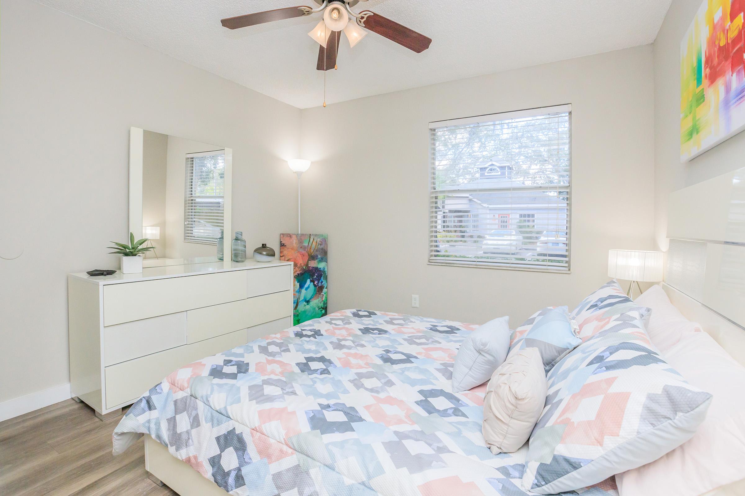 A cozy bedroom featuring a light-colored quilt with geometric patterns on a bed, a white dresser, and a mirror. The room has a ceiling fan, a floor lamp, and a colorful piece of wall art. Natural light shines through a window with blinds, creating a warm ambiance.