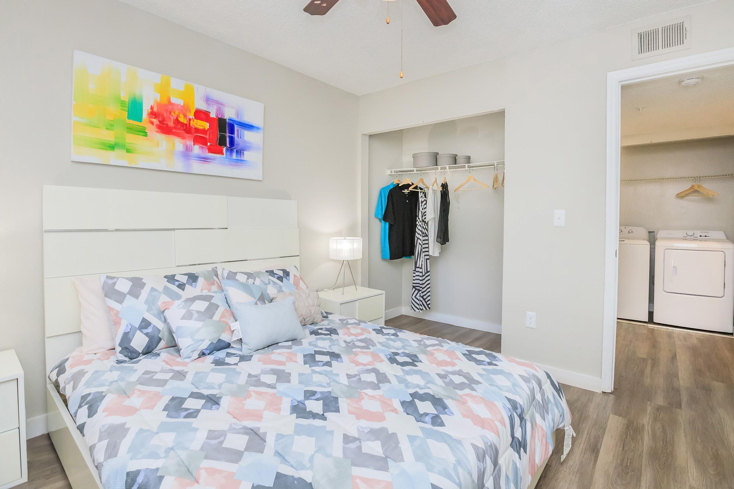 A modern bedroom featuring a patterned comforter on a bed, a bedside lamp, and colorful artwork on the wall. An open closet displays hanging clothes, while a separate laundry area is visible in the background. The room has light-colored walls and wooden flooring, creating a warm and inviting atmosphere.