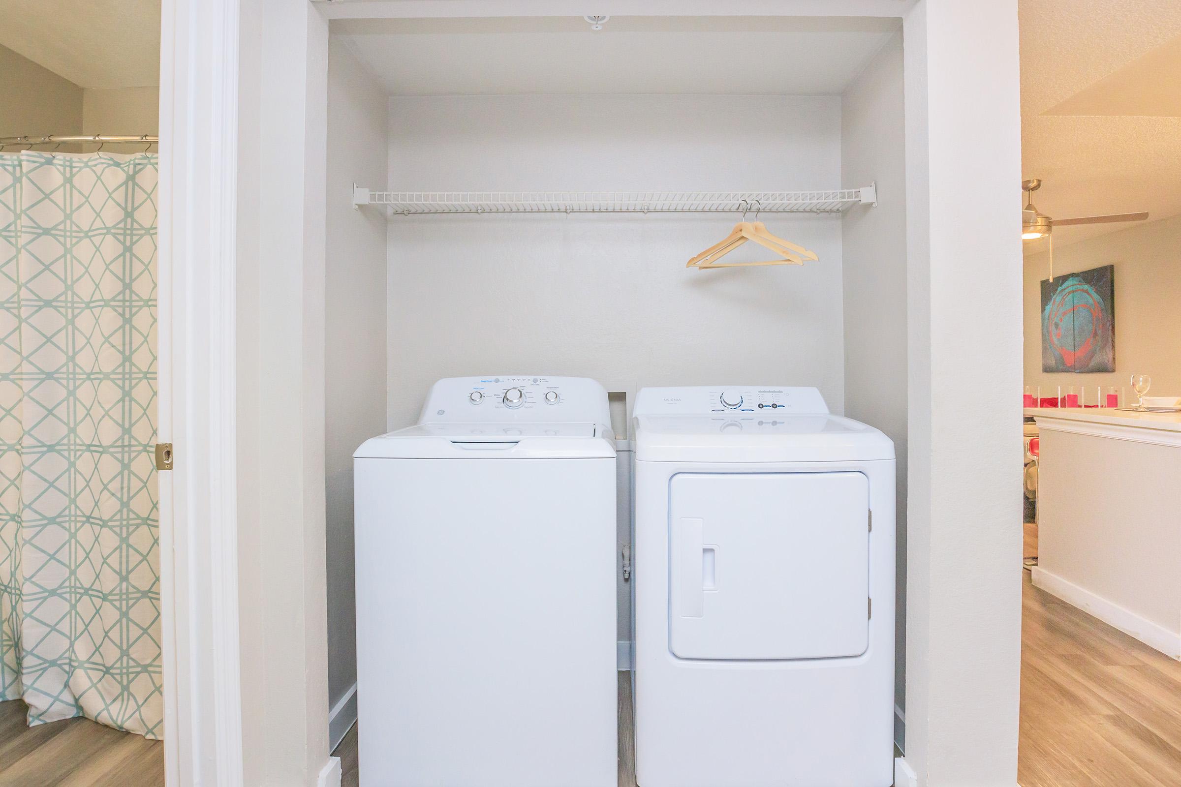 A compact laundry area featuring a washing machine and dryer side by side, with a wire shelf above. A wooden hanger is hanging on the shelf. In the background, a glimpse of a bathroom with a patterned shower curtain and a living area can be seen. The space is well-lit and modern.