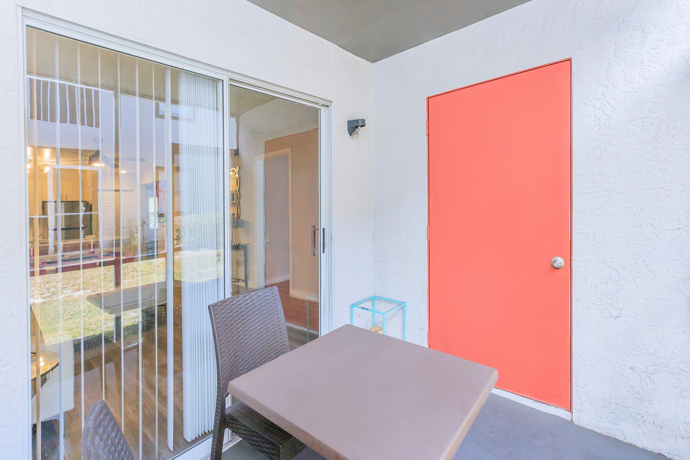 A small outdoor patio with a table and chairs, featuring a bright orange door. The sliding glass door leads into an interior space, showing a glimpse of a living area. Soft, natural light filters into the patio area, enhancing the contemporary design.