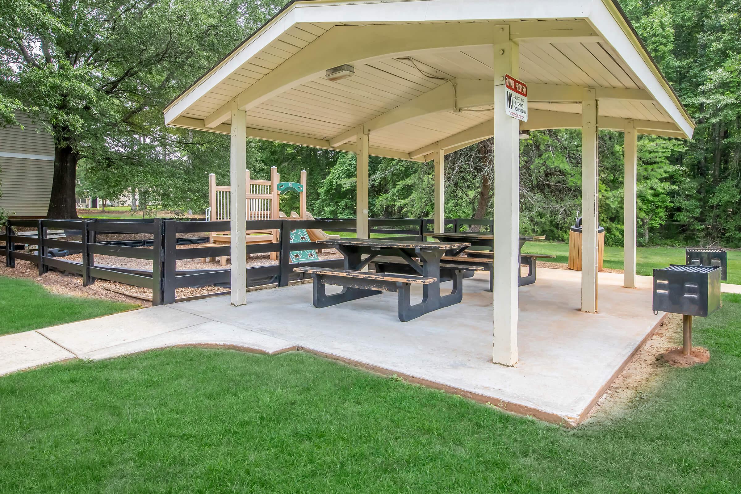 A covered pavilion with picnic tables set on a concrete surface, surrounded by lush green grass. In the background, there's a playground area with slides and climbing structures, enclosed by black wooden fencing. The pavilion provides shade for people enjoying outdoor activities.