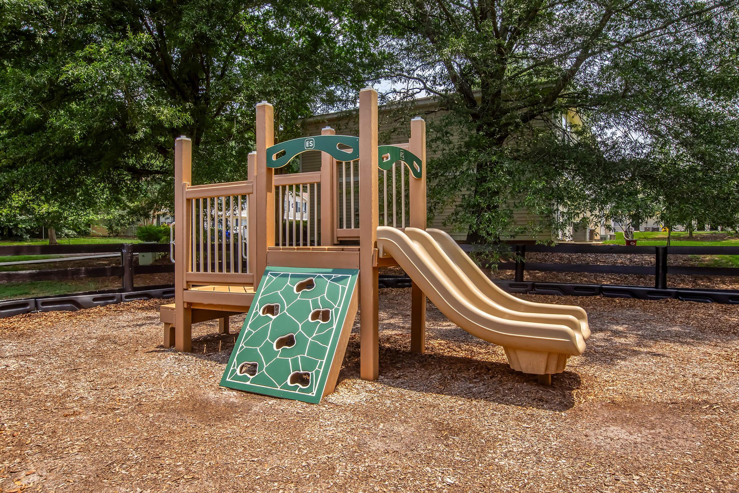 A wooden playground structure featuring a slide and an interactive leaf-themed panel with animal footprints. The area is surrounded by mulch and trees, creating a natural play environment.