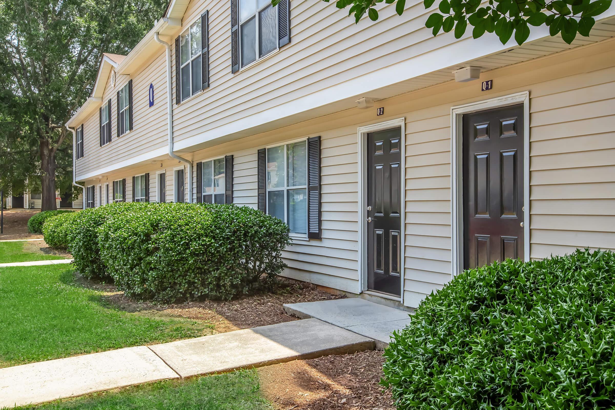 Two-story apartment building with beige siding, featuring multiple windows and black doors numbered 13 and 14. Neatly trimmed bushes line the walkway leading to the doors, with a well-maintained lawn in front. Trees are visible in the background, contributing to a pleasant residential setting.