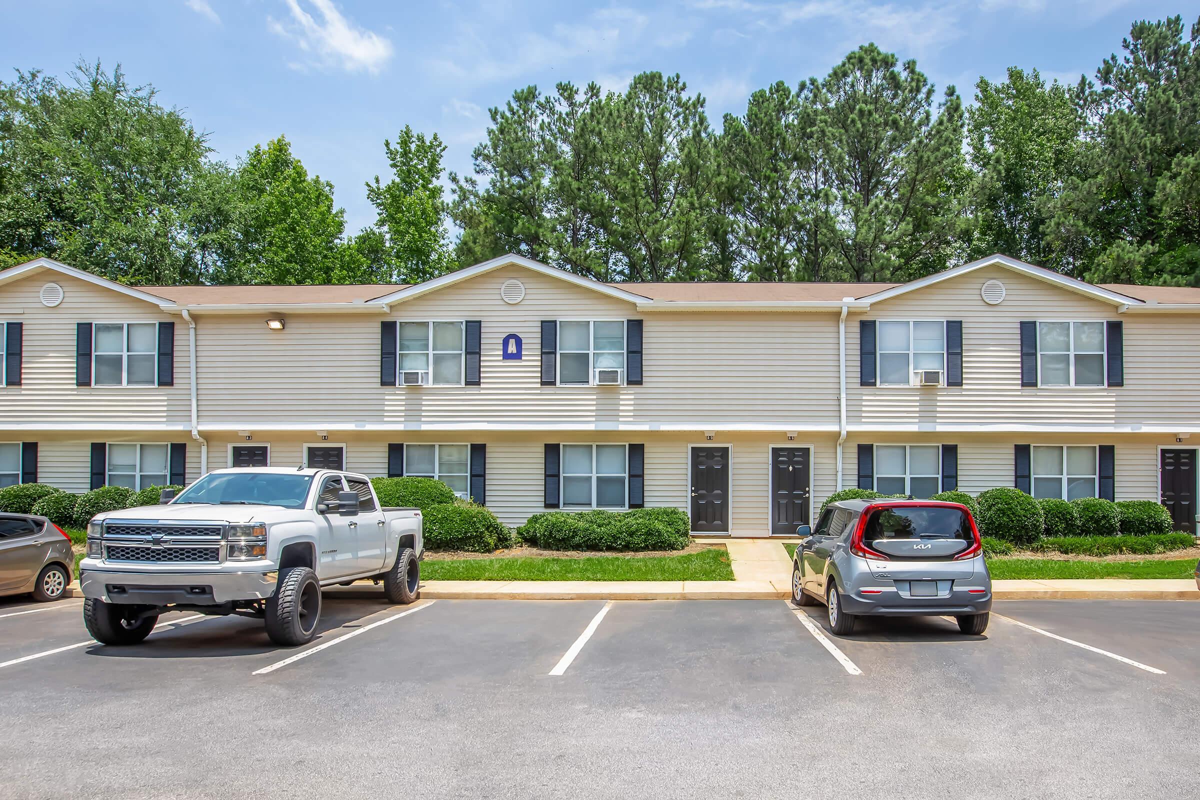 A row of two-story apartment buildings with multiple units visible. Two parked vehicles are in the foreground: a gray pickup truck and a small red SUV. The lawn is neatly trimmed with shrubs, and trees line the background under a clear blue sky.