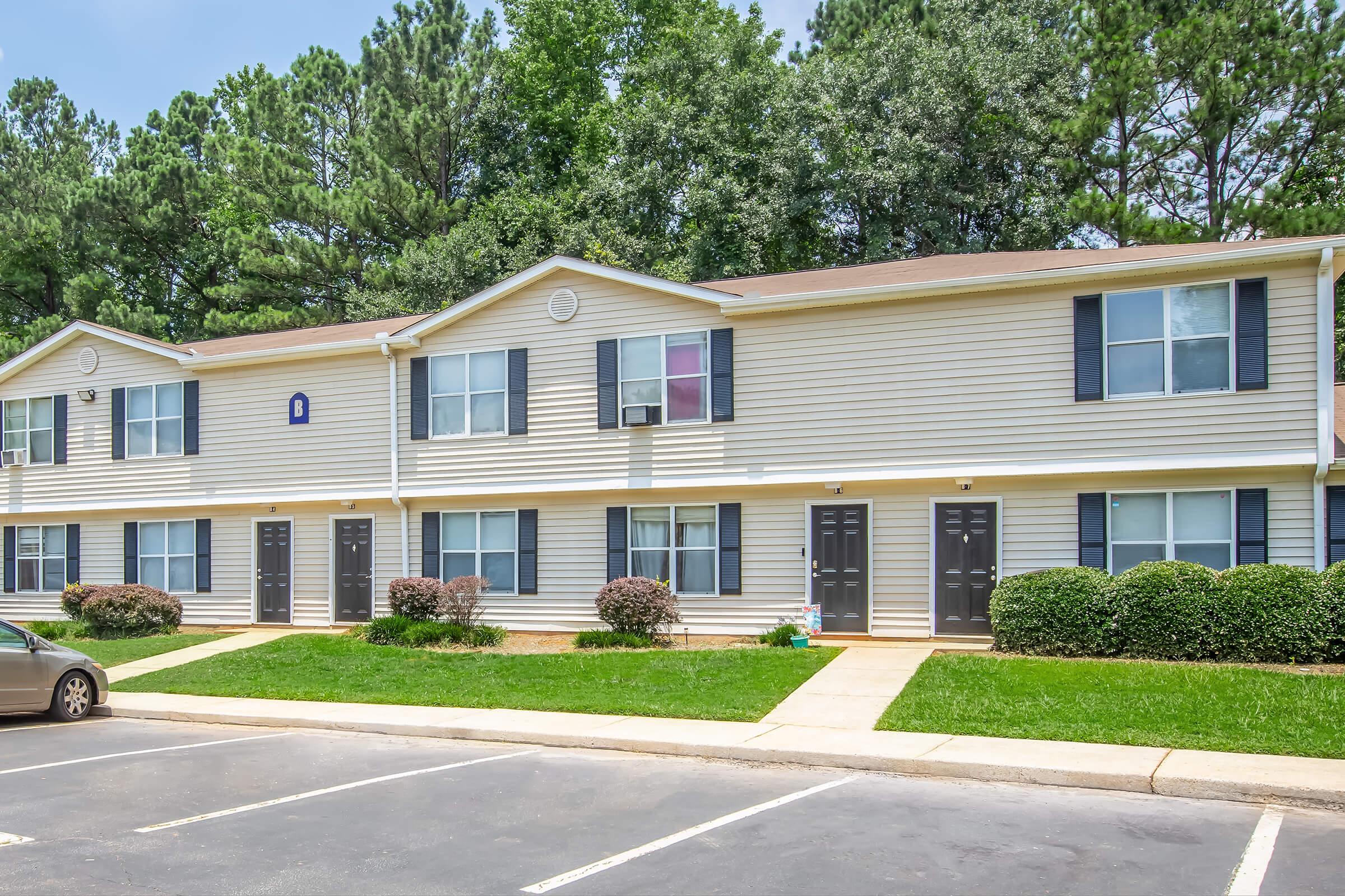 A two-story apartment building with light grey siding, dark blue doors, and windows. The surrounding area features neatly trimmed grass and shrubs. In front, there are parking spaces with a few cars parked. Tall green trees are visible in the background under a clear blue sky.