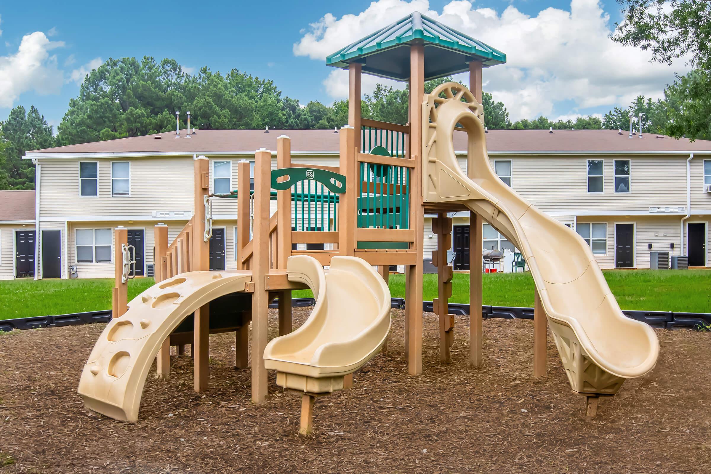 Playground equipment featuring two slides, a climbing structure, and a green roof. The setup is situated on a bed of mulch, surrounded by grass and apartment buildings in the background under a partly cloudy sky.