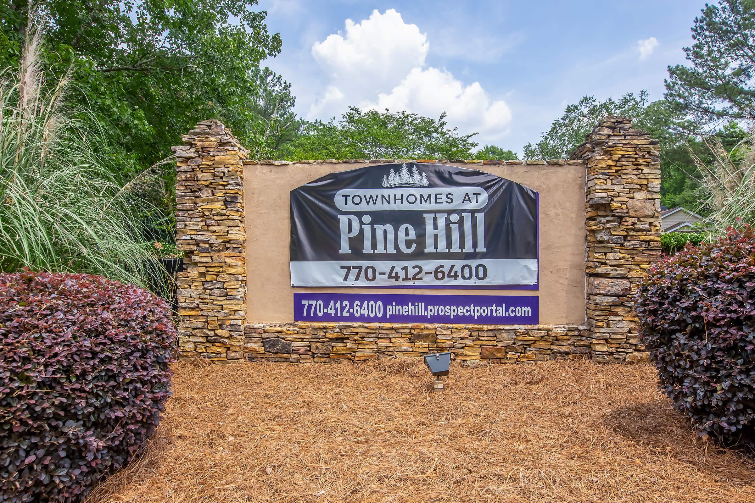 Sign for "Townhomes at Pine Hill" featuring a stone structure, with landscaping including shrubs and grass. The sign includes a phone number (770-412-6400) and a website (pinehill.prospectportal.com), set against a backdrop of blue skies and trees.