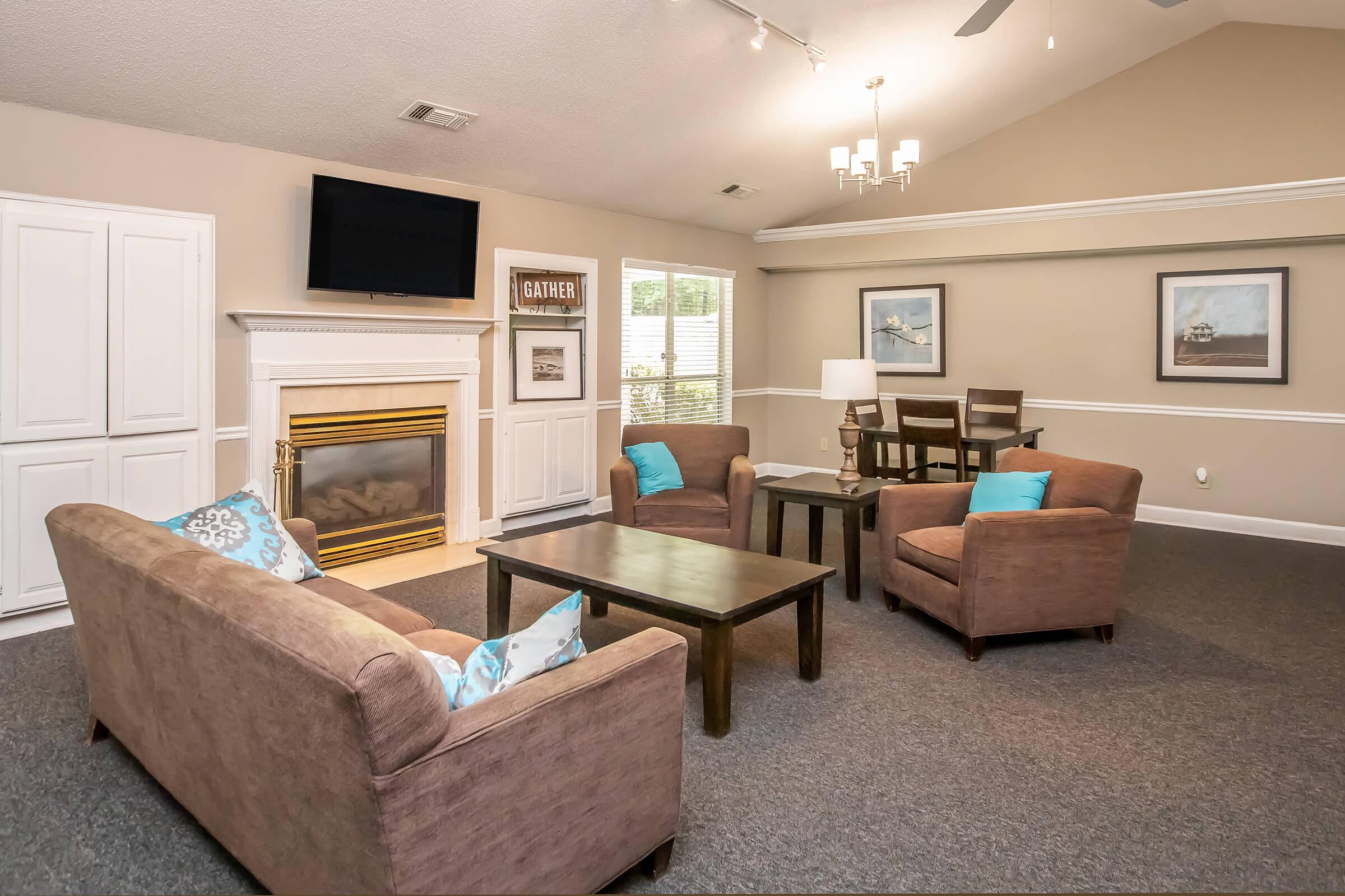 Cozy living area featuring a comfortable seating arrangement with two armchairs and a sofa adorned with blue cushions, a wooden coffee table, and a wall-mounted television. A fireplace is positioned beneath a decorative mantle, complemented by framed artwork and a dining table in the background.