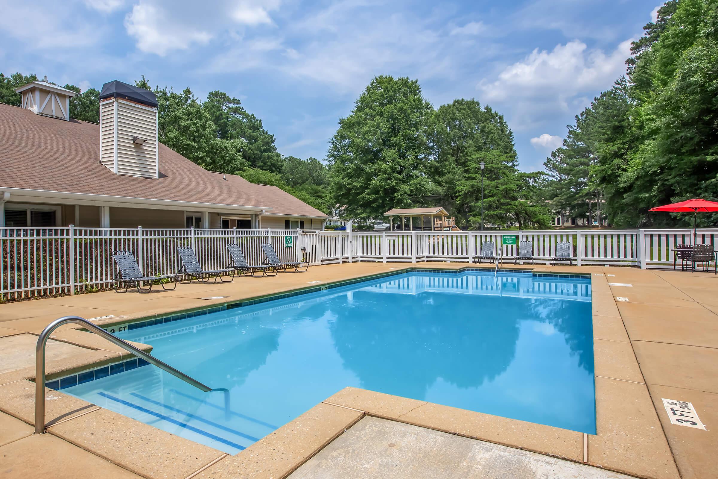 A clear blue swimming pool surrounded by a tan deck, with lounge chairs positioned nearby. A white fence encloses the area, and green trees provide a natural backdrop. The sky is partly cloudy, creating a bright and inviting atmosphere.