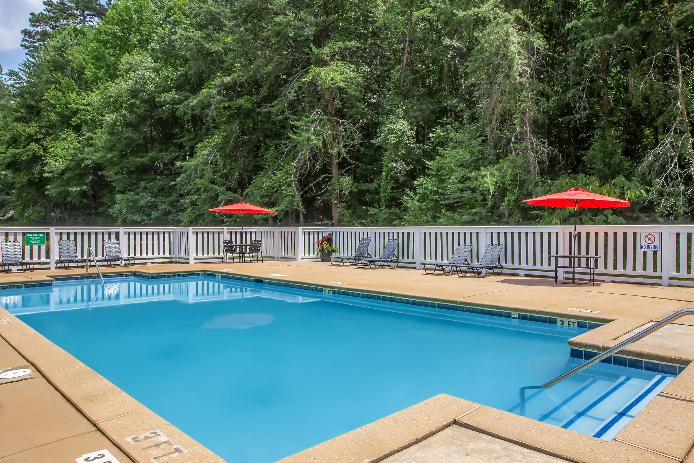 A clear outdoor swimming pool surrounded by a tan deck, with several lounge chairs arranged nearby. Bright red umbrellas provide shade, and lush green trees line the background. A white fence encloses the pool area, with signs indicating rules for safety. The scene is inviting and well-maintained.