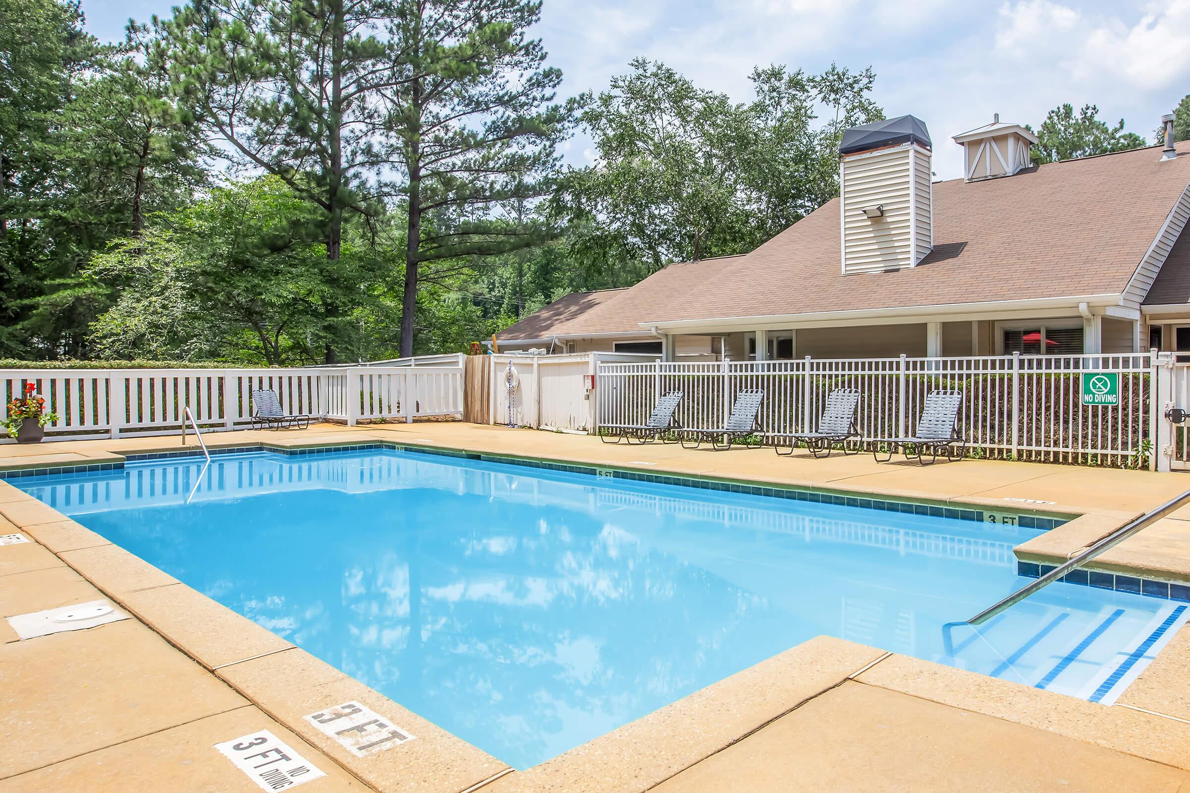 A clear blue swimming pool surrounded by a concrete deck with several lounge chairs. Green trees and a building are visible in the background, creating a relaxing outdoor atmosphere. The pool area features safety markings, including depth indicators.