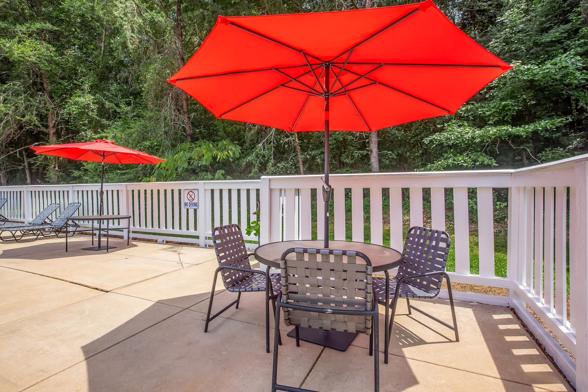 A serene outdoor patio area featuring a round table with chairs, complemented by two large red umbrellas for shade. The setting includes a white railing and lush greenery in the background, creating a relaxing atmosphere perfect for outdoor gatherings.