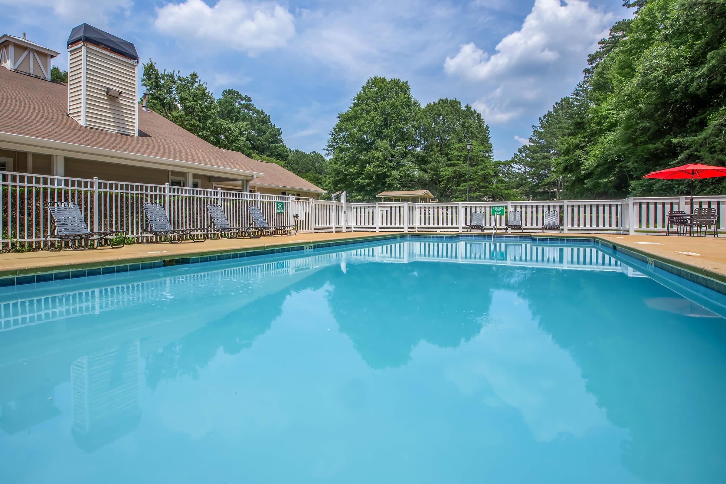 A clear blue swimming pool reflects the surrounding greenery and a partly cloudy sky. In the background, there is a building with a brown roof and white railing. Lounge chairs are lined up beside the pool, with a red umbrella providing shade nearby. The scene conveys a relaxing outdoor atmosphere.