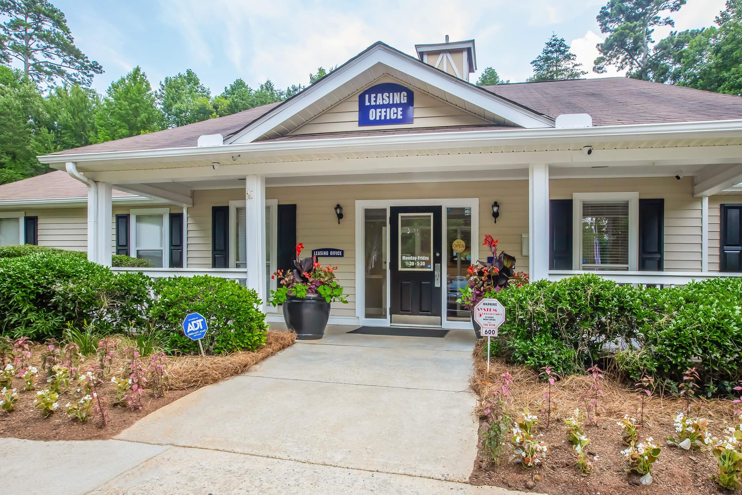 A welcoming leasing office with a covered entrance, surrounded by well-maintained flower beds and shrubs. The building features a dark door, light-colored siding, and a sign indicating "Leasing Office," set against a backdrop of trees and blue sky.