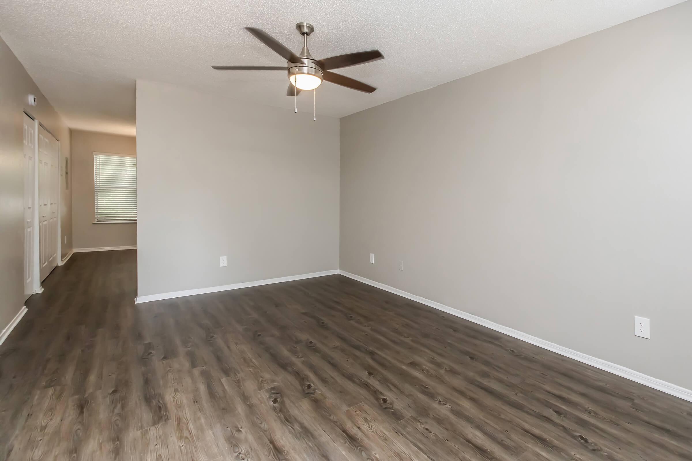 A spacious, empty living room featuring a ceiling fan and light fixture. The hardwood-style flooring is dark, and the walls are painted in a light gray color. A hallway is visible in the background, leading to another room with windows. The space is bright and ready for furniture arrangement.