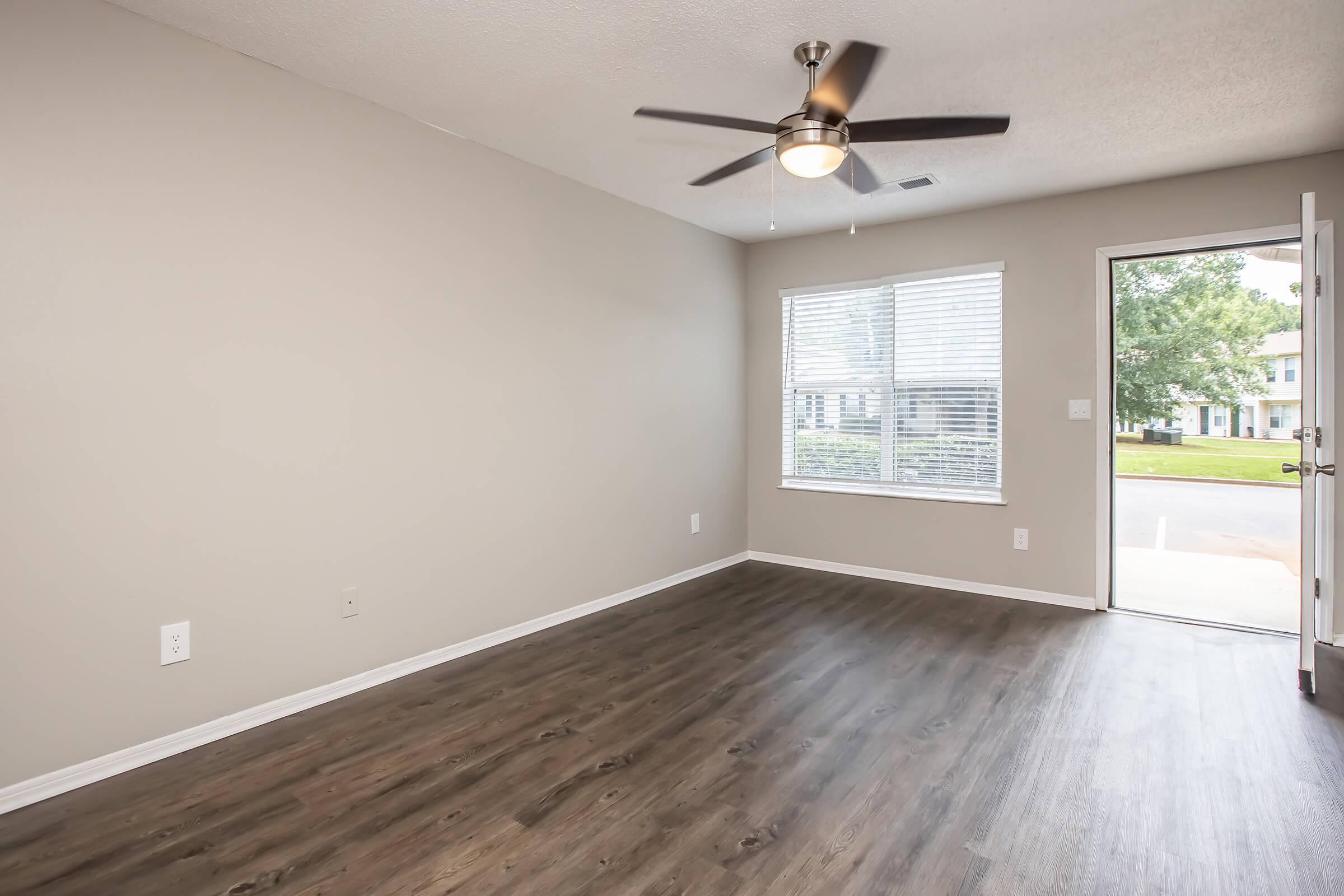 Empty interior of a room featuring a ceiling fan, a large window with blinds, and an open door leading outside. The walls are painted light gray, and the floor is covered in wood-like laminate. Natural light fills the space, and the overall atmosphere is bright and inviting.