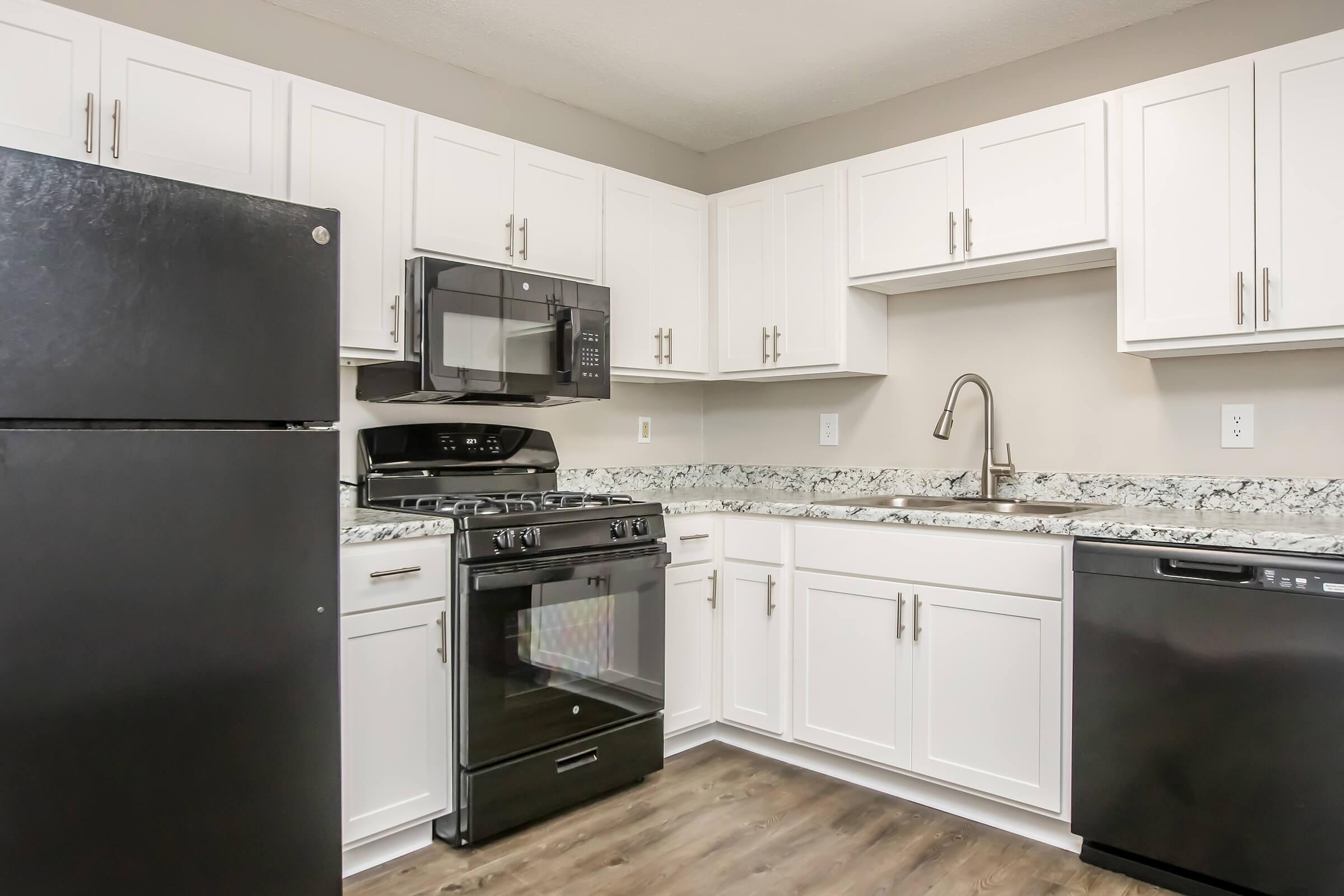 A modern kitchen featuring white cabinetry, granite countertops, and stainless steel appliances. It includes a black refrigerator, a gas stove with a microwave above, and a black dishwasher. The flooring is dark wood, creating a sleek and contemporary look.